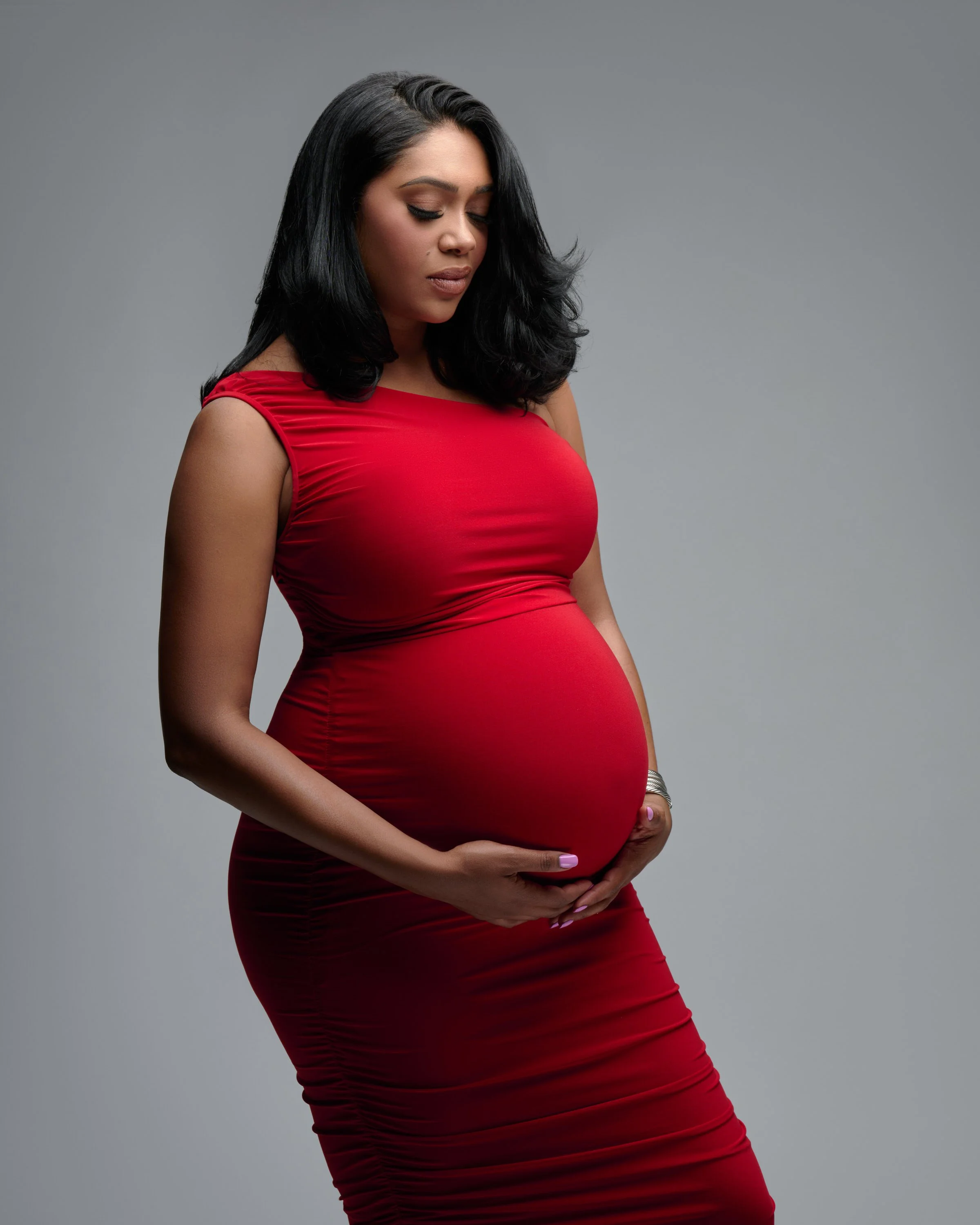 A pregnant woman with dark hair in a red dress holds her belly and looks downward against a plain grey background.