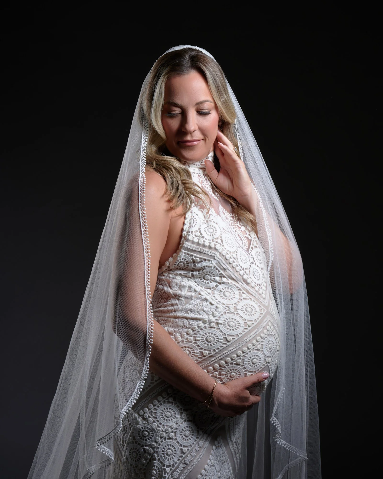Pregnant mother in her wedding dress and veil holding her bump during a Denver studio session.