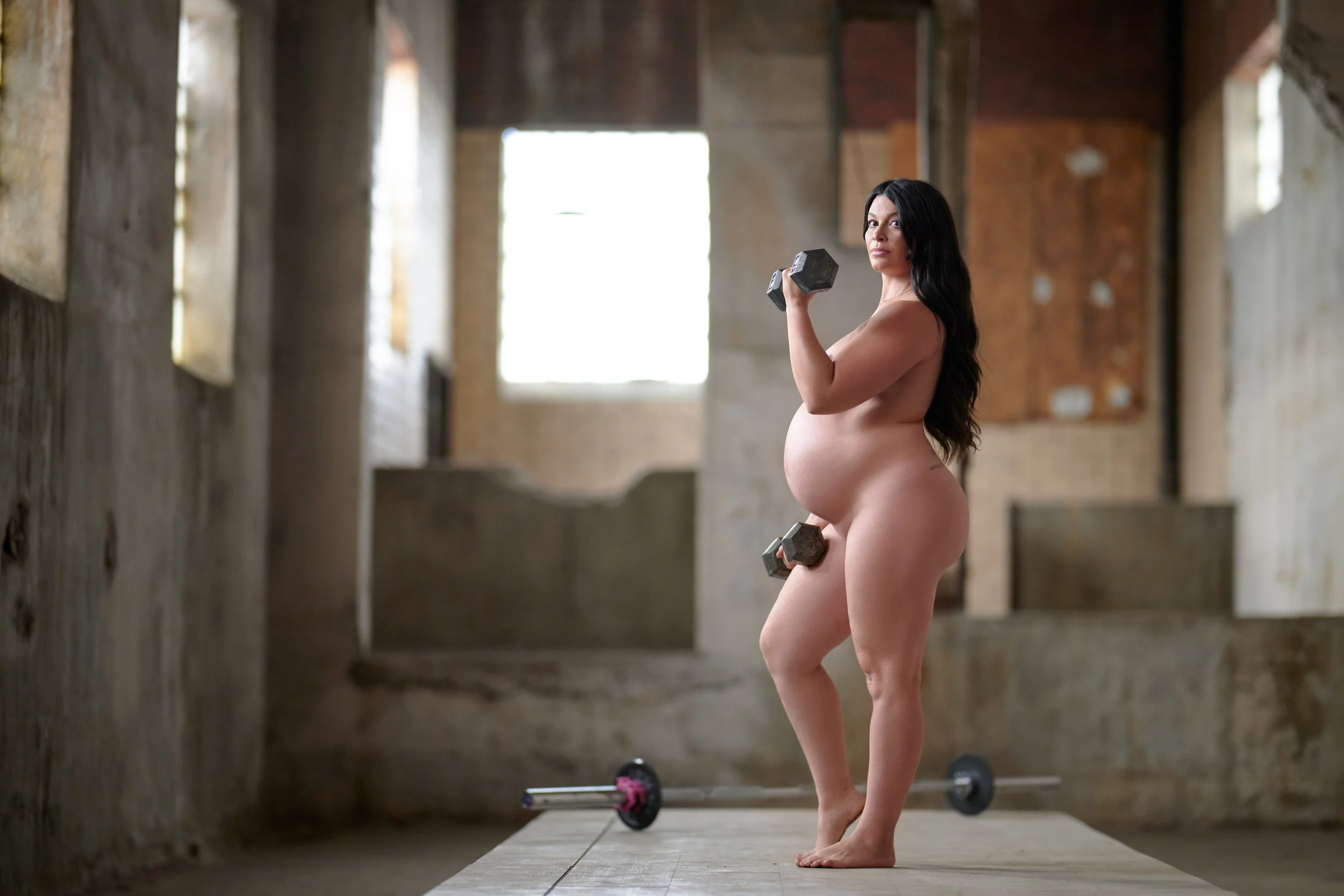 Dumbbell curl in a bare warehouse, window light raking across the bump.