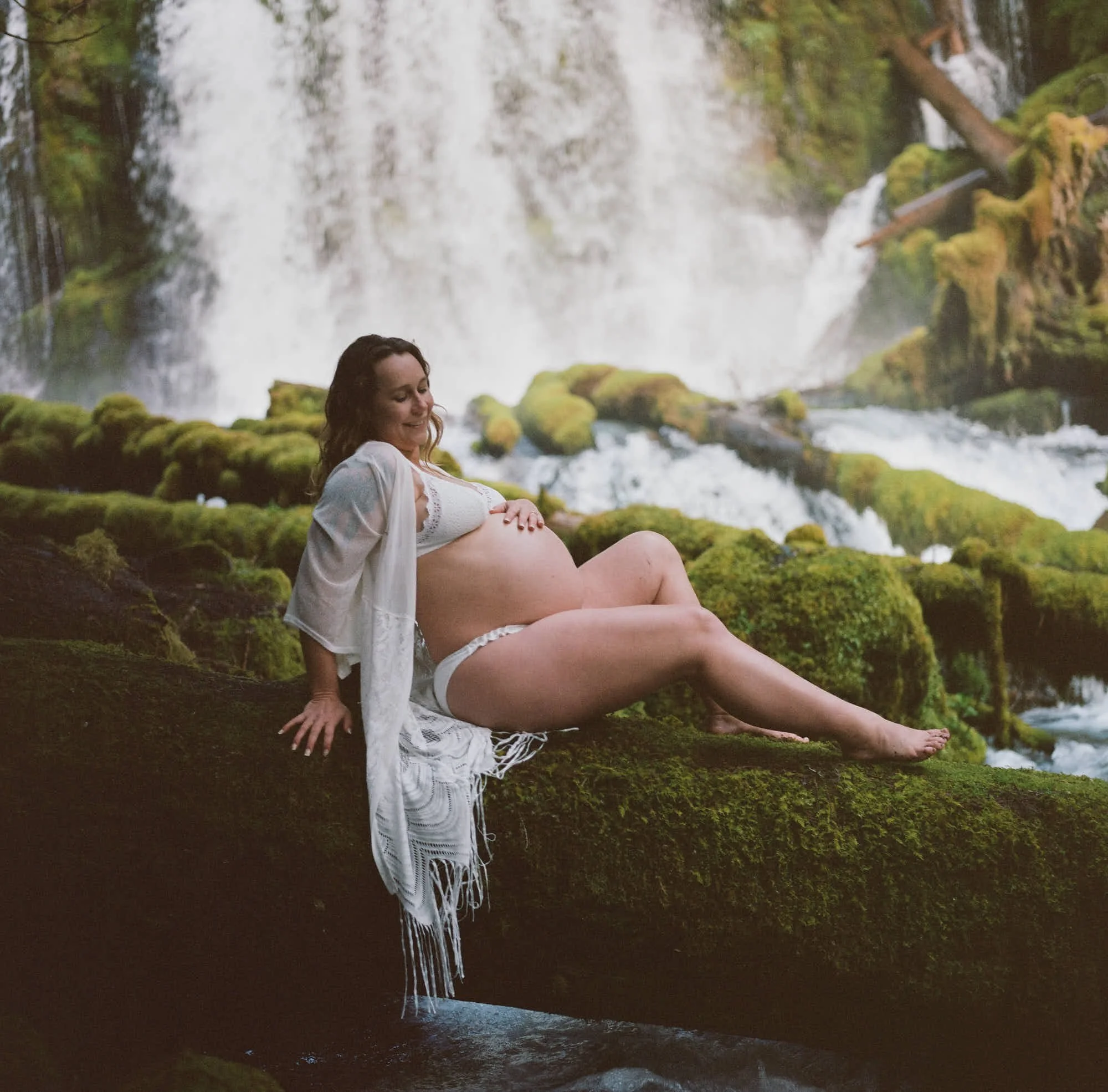 Seated on a moss-covered log in white lace, waterfalls pouring down the cliff behind.