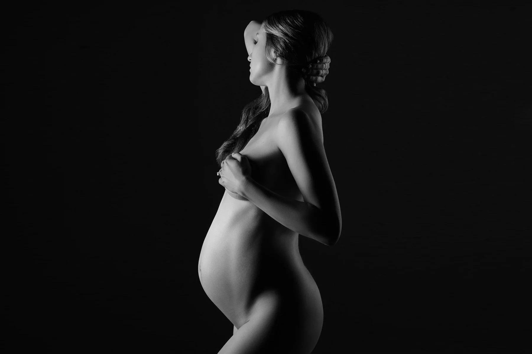 Profile in soft light against black, hands at her chest, a black and white bare form study.