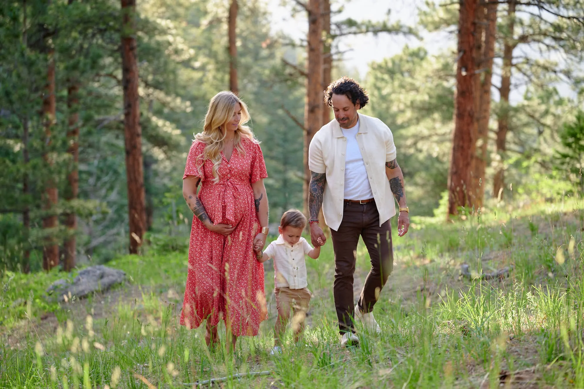 Parents walk hand in hand with their son through tall pines, red florals and warm light.