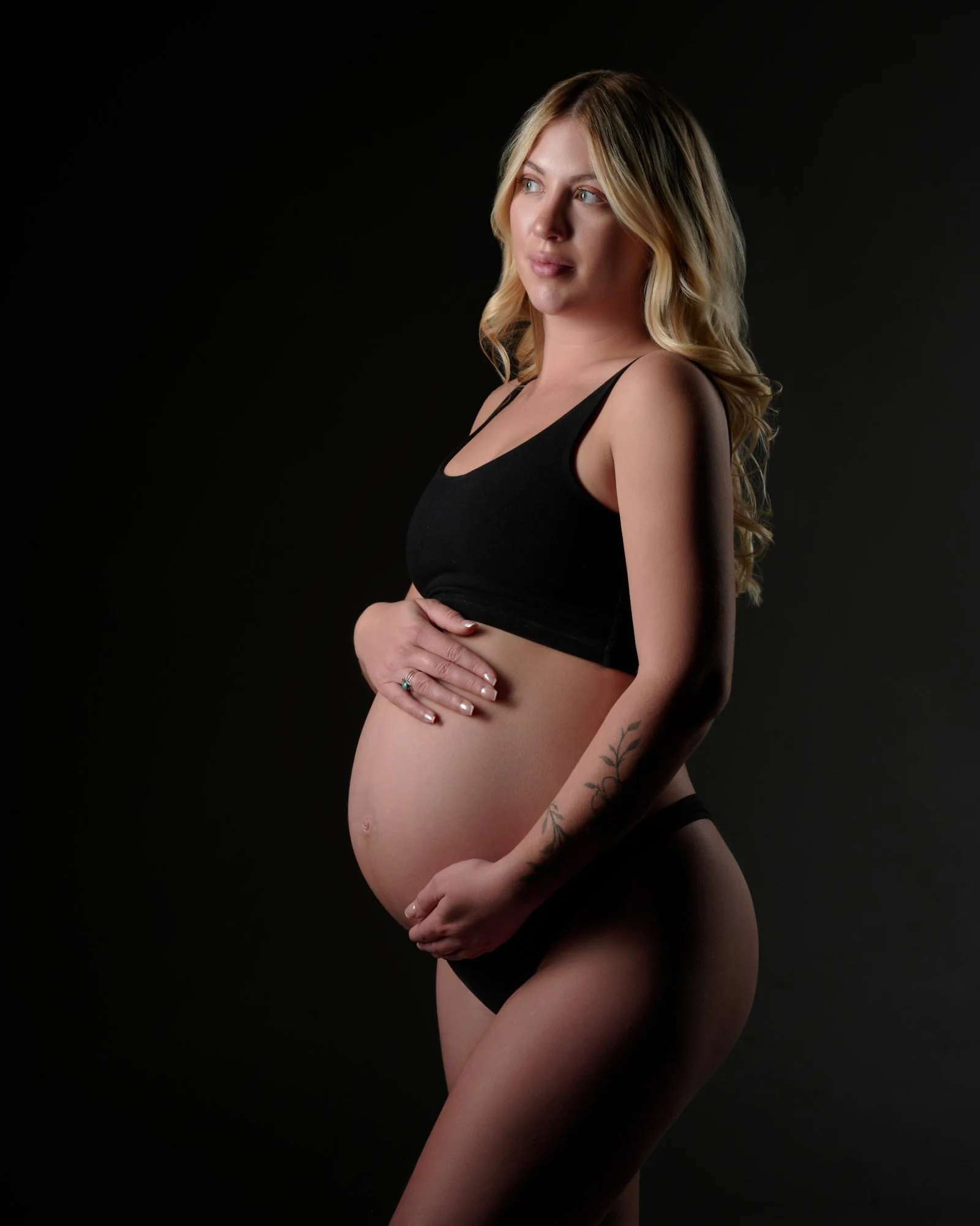 Studio maternity photoshoot pose in a black two-piece, both hands cradling the bare bump against a dark backdrop.