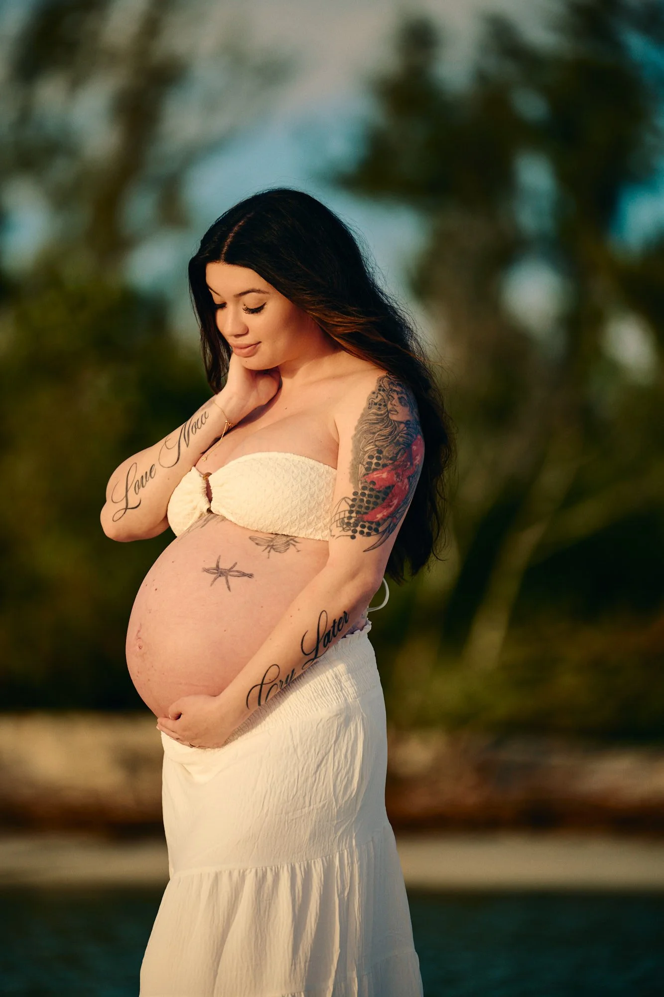 White top and tiered skirt beside mangroves, tattoos framing the bump in warm light.