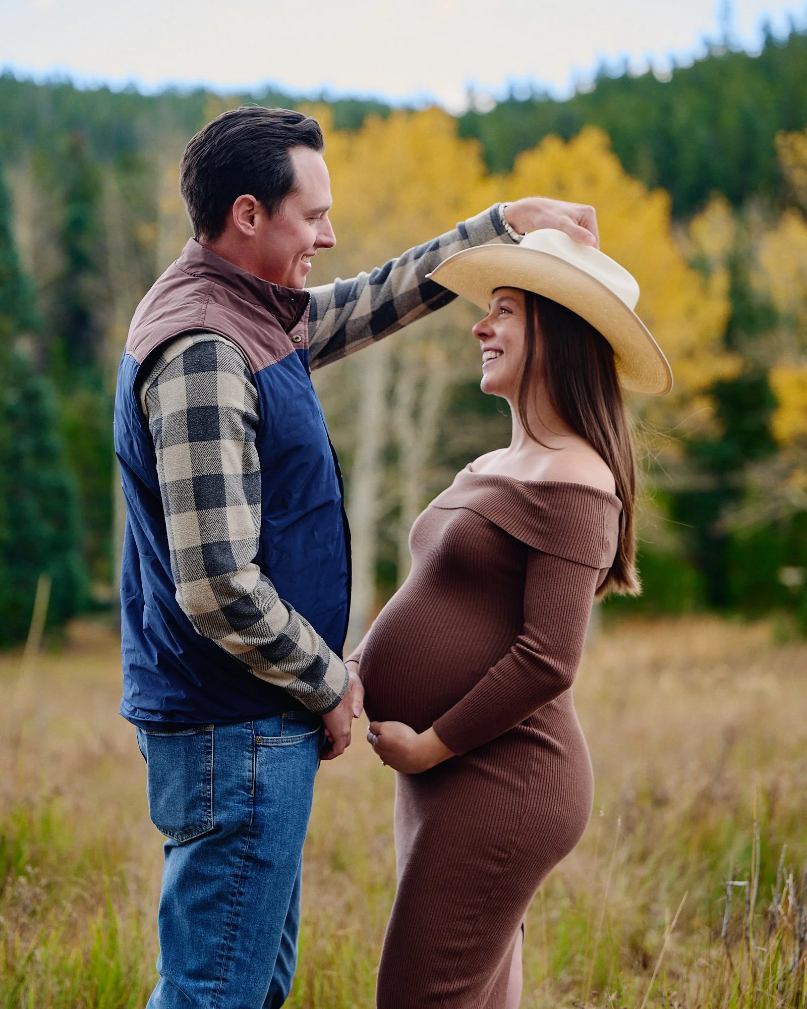 Outdoor fall maternity photoshoot in a brown dress, her partner setting a wide-brim hat as gold aspens turn behind them.