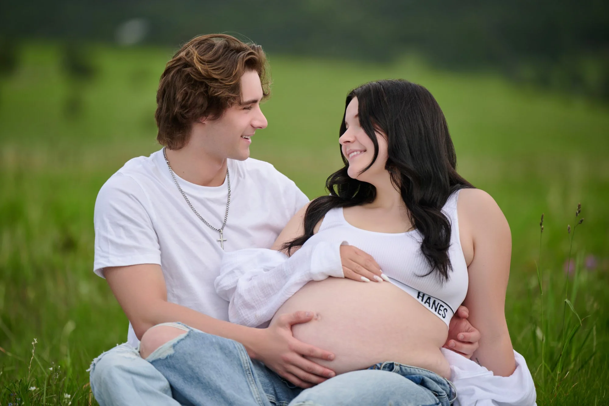 A young couple sitting outdoors in a grassy field, with the woman pregnant, smiling at each other.