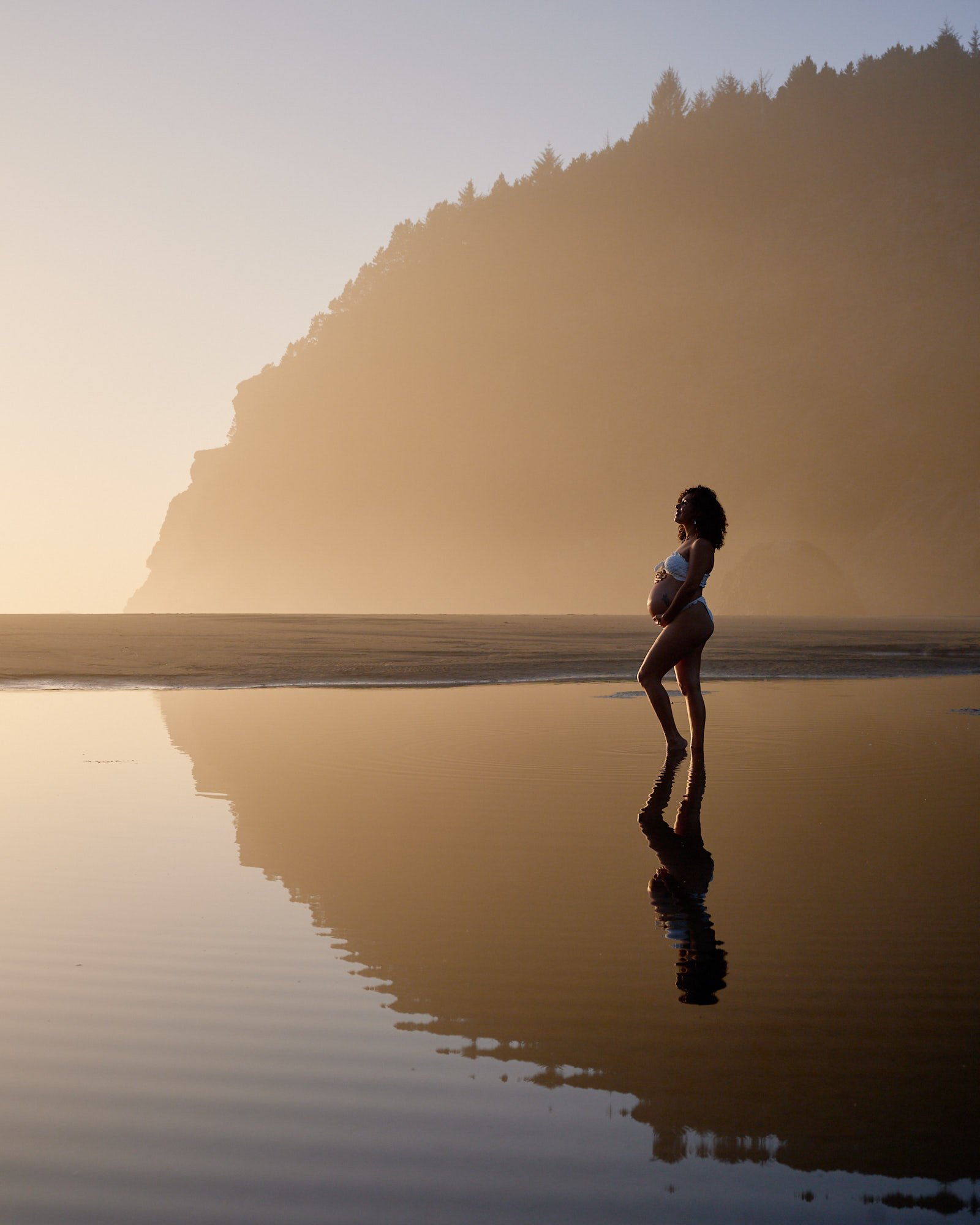 Walking the wet sand at low tide, her reflection below and a headland fading into the haze.
