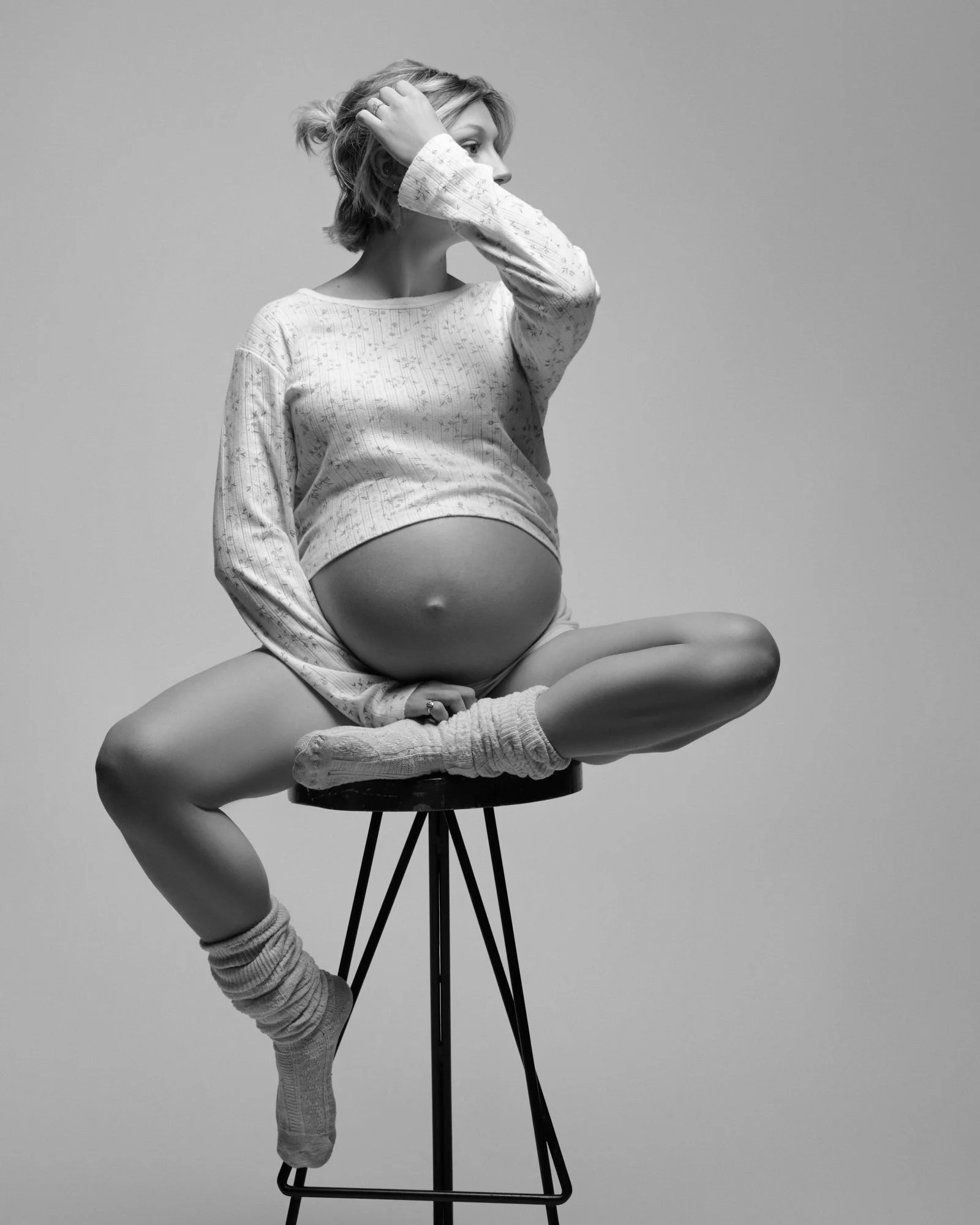 Mother-to-be sits cross-legged on a tall stool in a floral top, relaxed and candid.