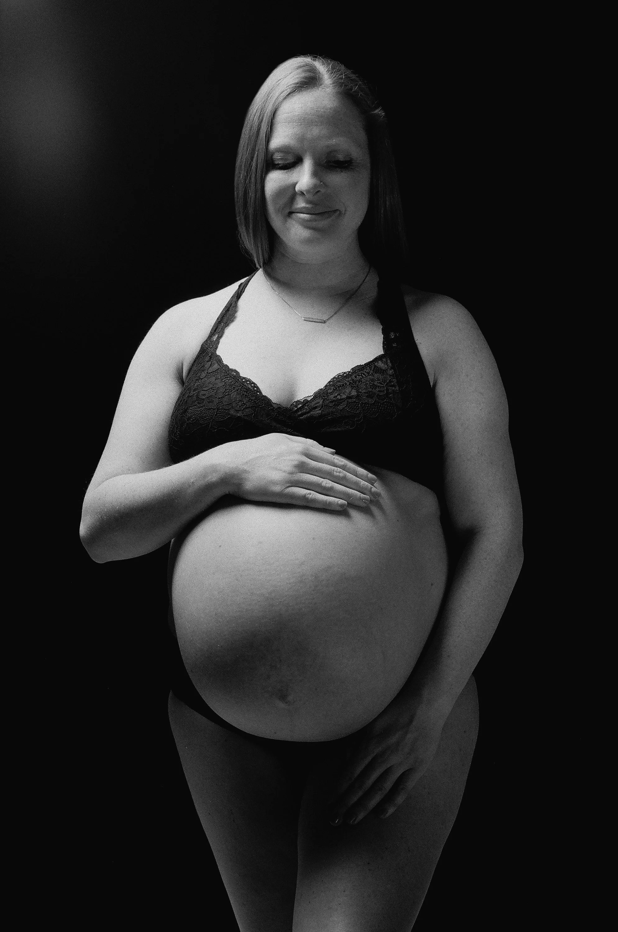 Black lace and low key light, both hands cradling the bump, deep shadow behind her on film.