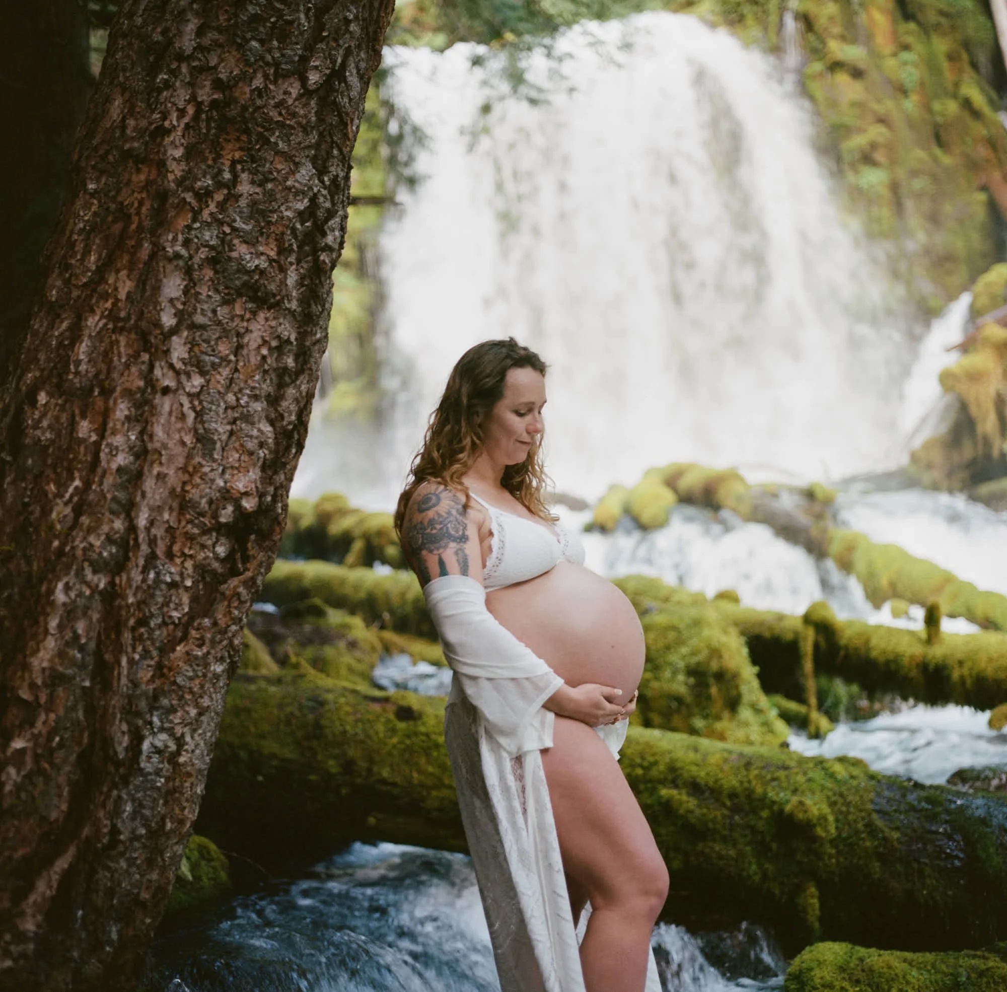 Tattooed arm and bare bump beside a fir trunk, waterfall spilling over the rock behind.