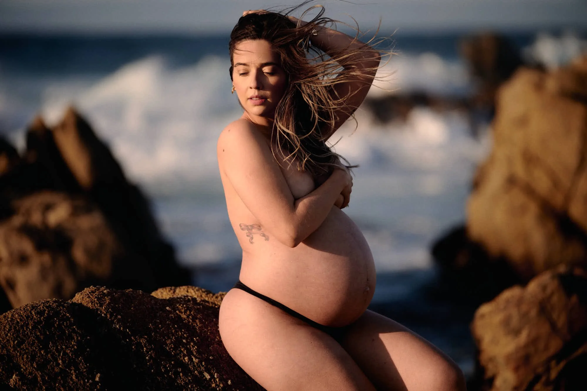 Seated on shoreline rocks with hair blowing across her face, surf breaking white behind, bare form.