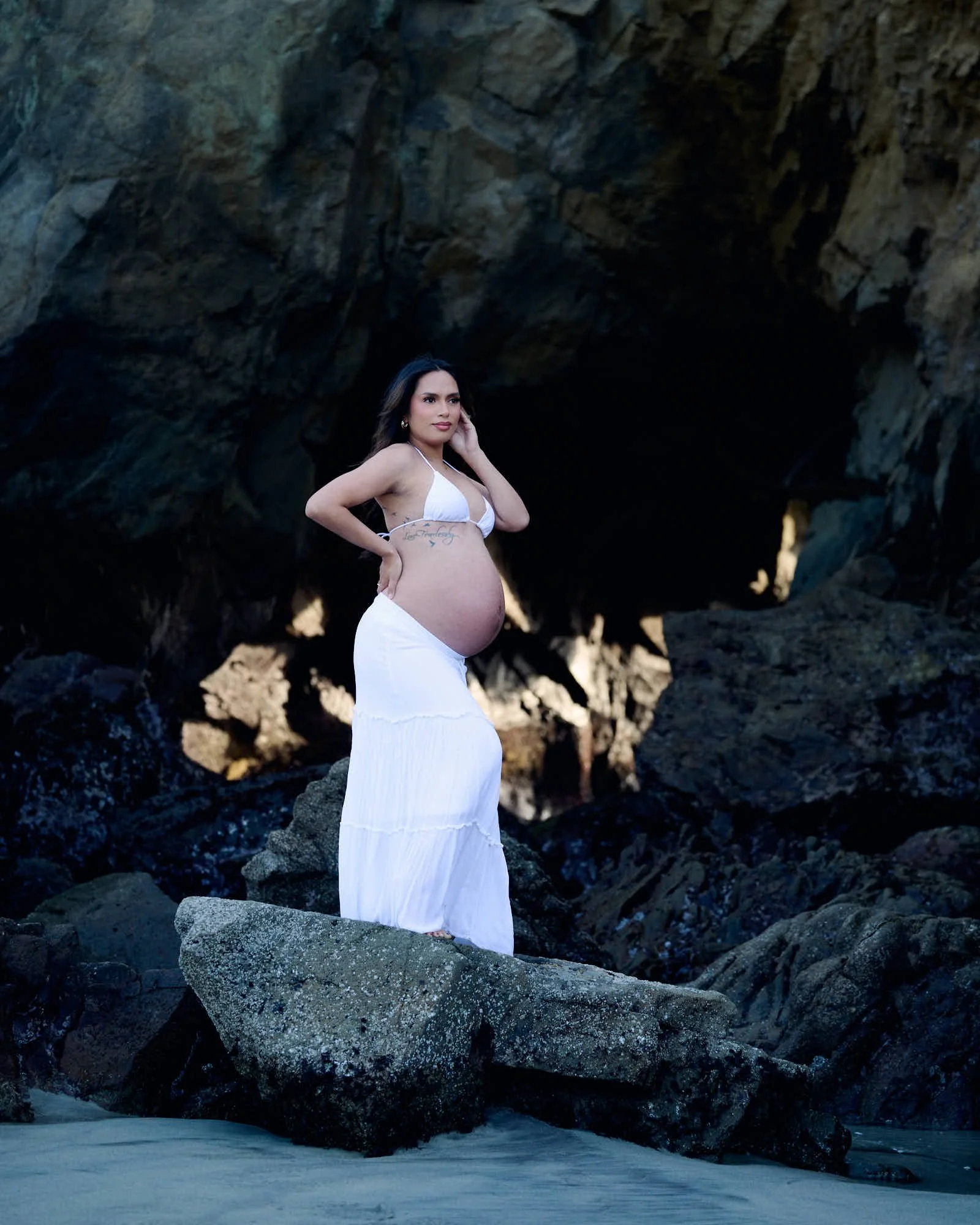 White two-piece on a boulder inside a sea cave, dark rock walls framing the Big Sur coastline.