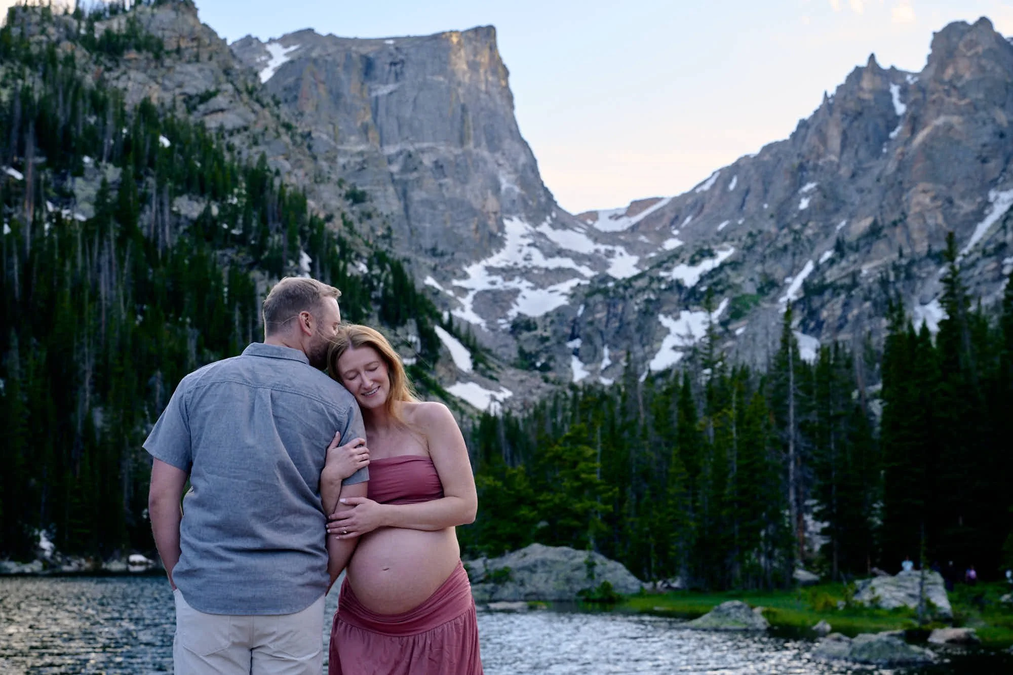 Partner kisses her forehead at the edge of an alpine lake at dusk, snow-streaked peaks behind, rose two-piece over the bump.