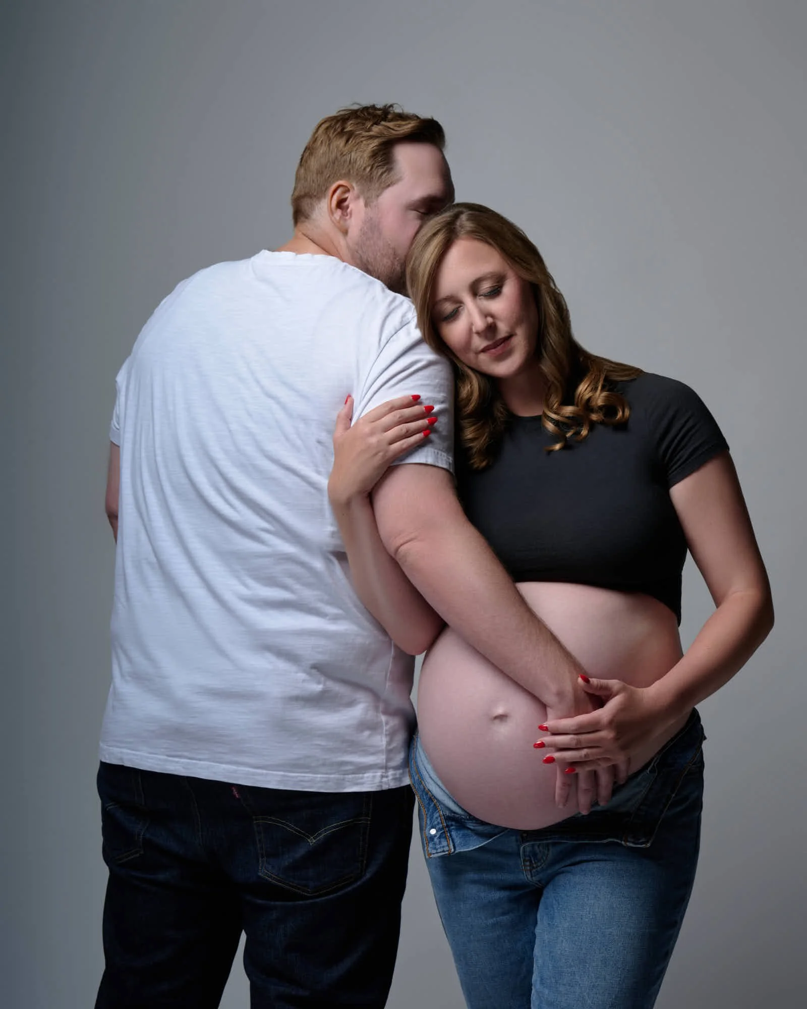 Partner kisses her forehead, a black crop top and unbuttoned jeans framing the bump.