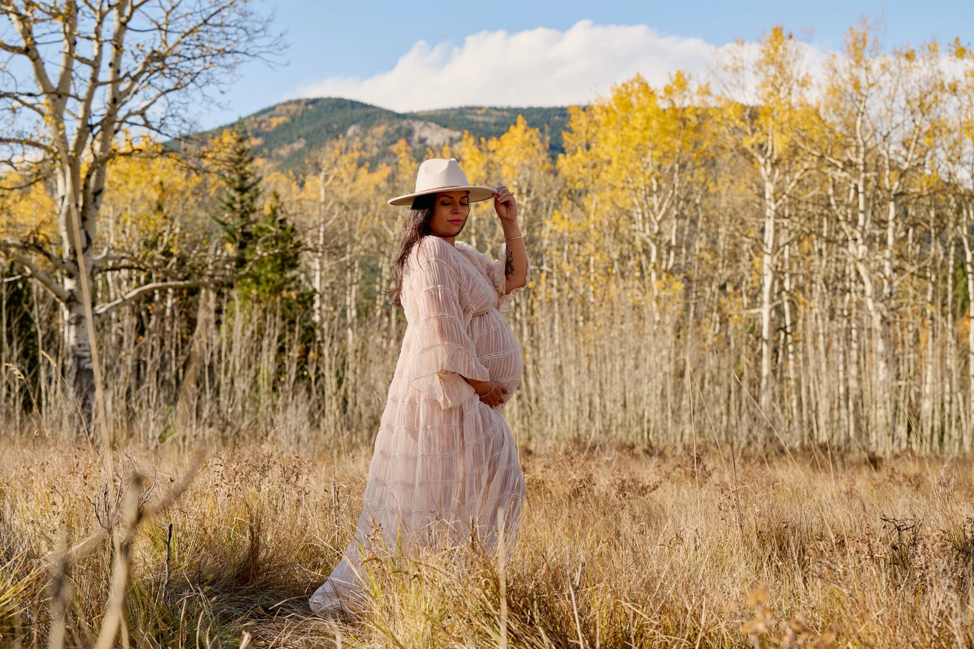 Expecting mother in a cream dress and wide-brim hat among golden aspens during a fall maternity session in Colorado.
