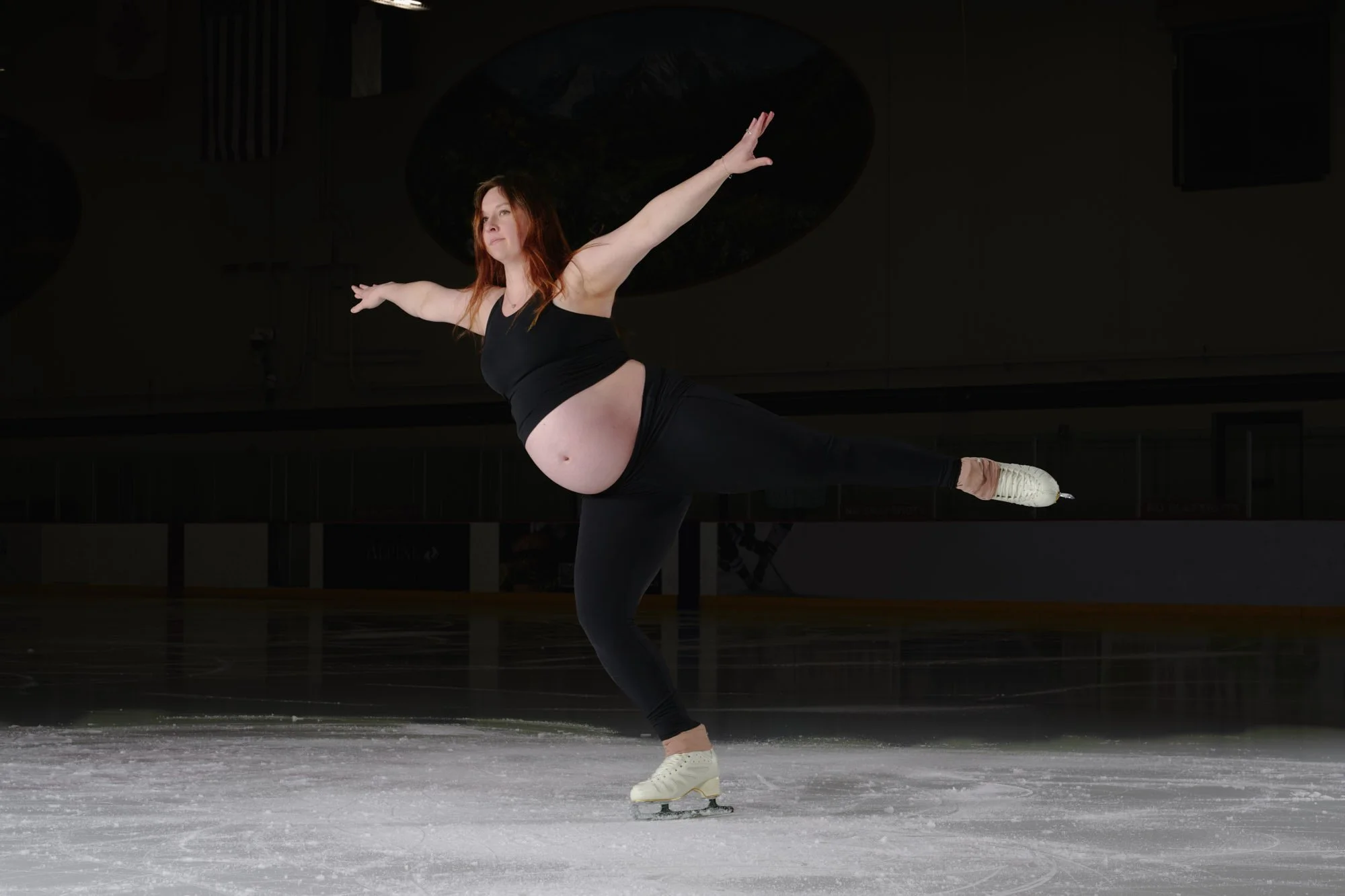Figure skater in an arabesque on the ice, arms out and the bump bare under a black crop.
