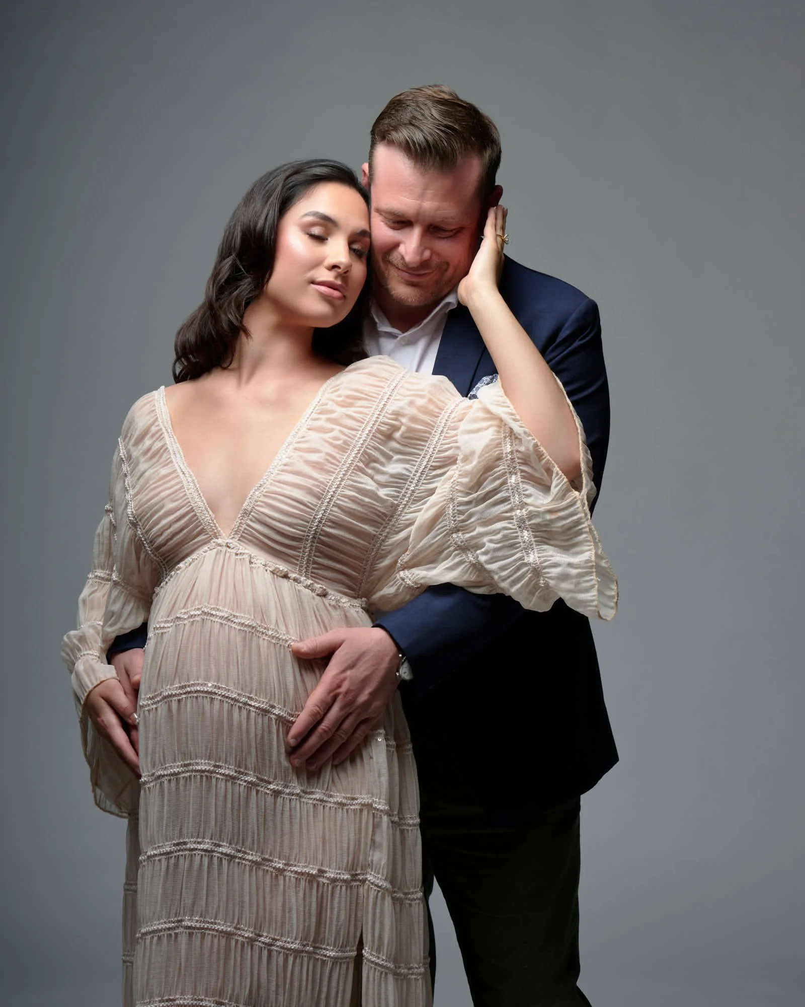 Sheer cream gown and a navy suit, partner embracing the baby bump from behind.