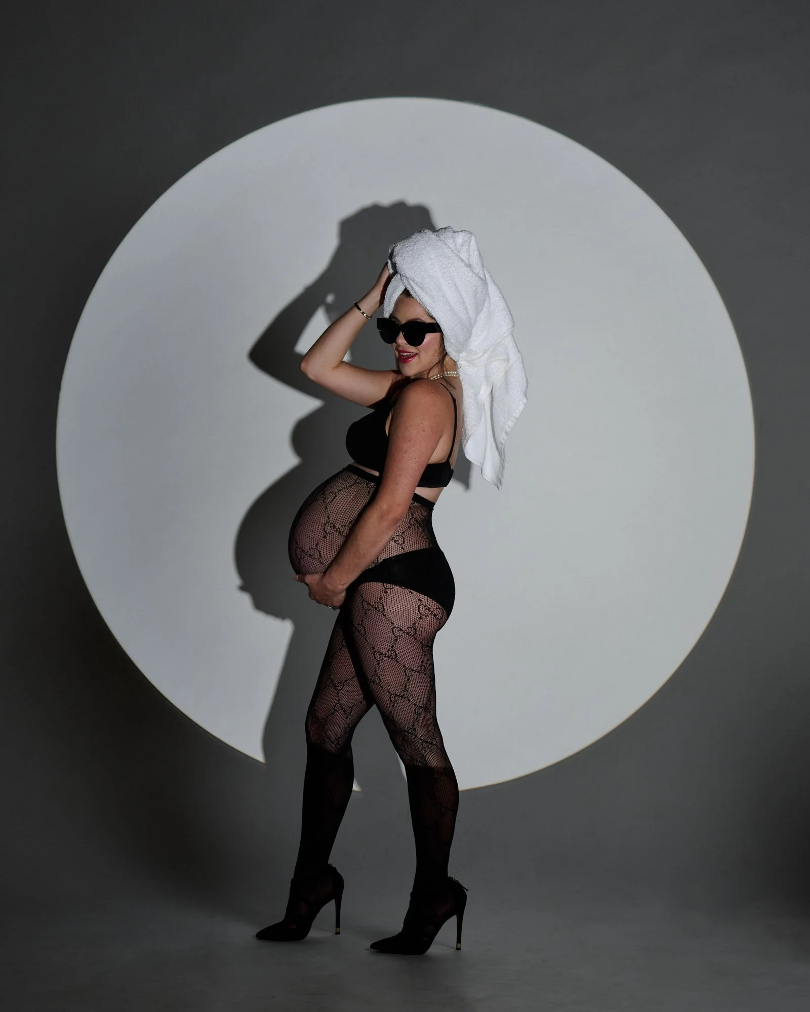 A pregnant woman in black lingerie, fishnet stockings, and high heels, wearing sunglasses, with a towel wrapped around her head, stands against a plain background with a circular spotlight, striking a pose and smiling.