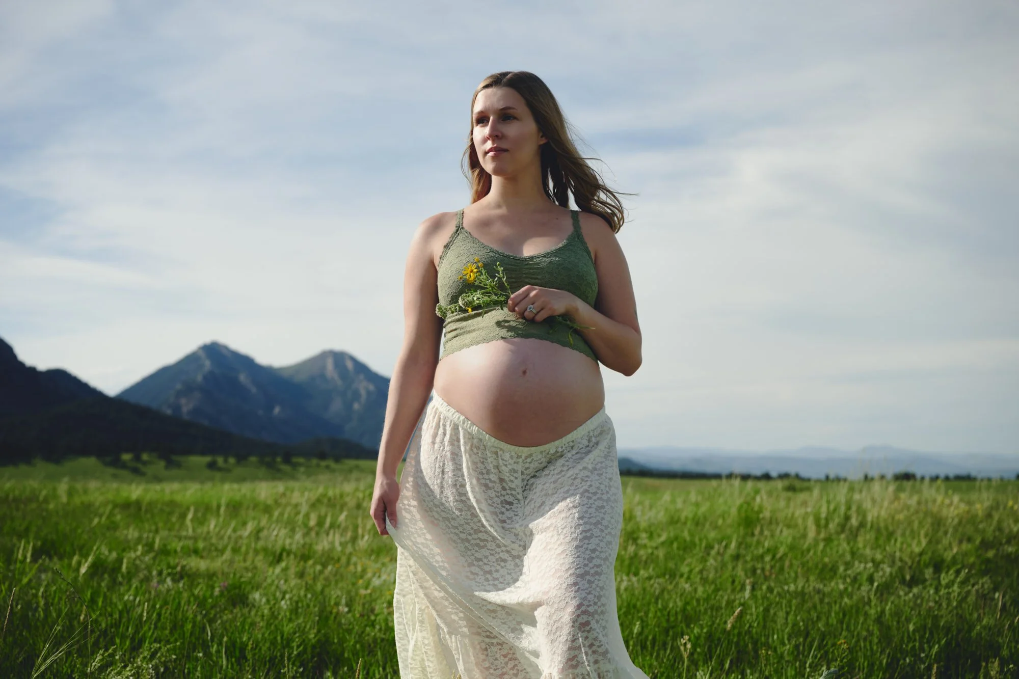 Expecting mother in a green tank top and white lace skirt standing in a summer meadow below Colorado mountains.