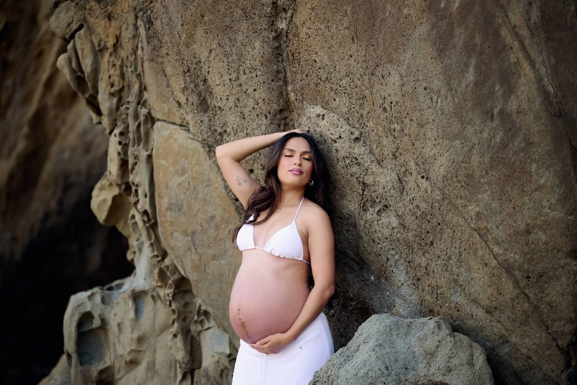 White lace top and skirt against a weathered rock face, eyes closed in the Monterey coast light.