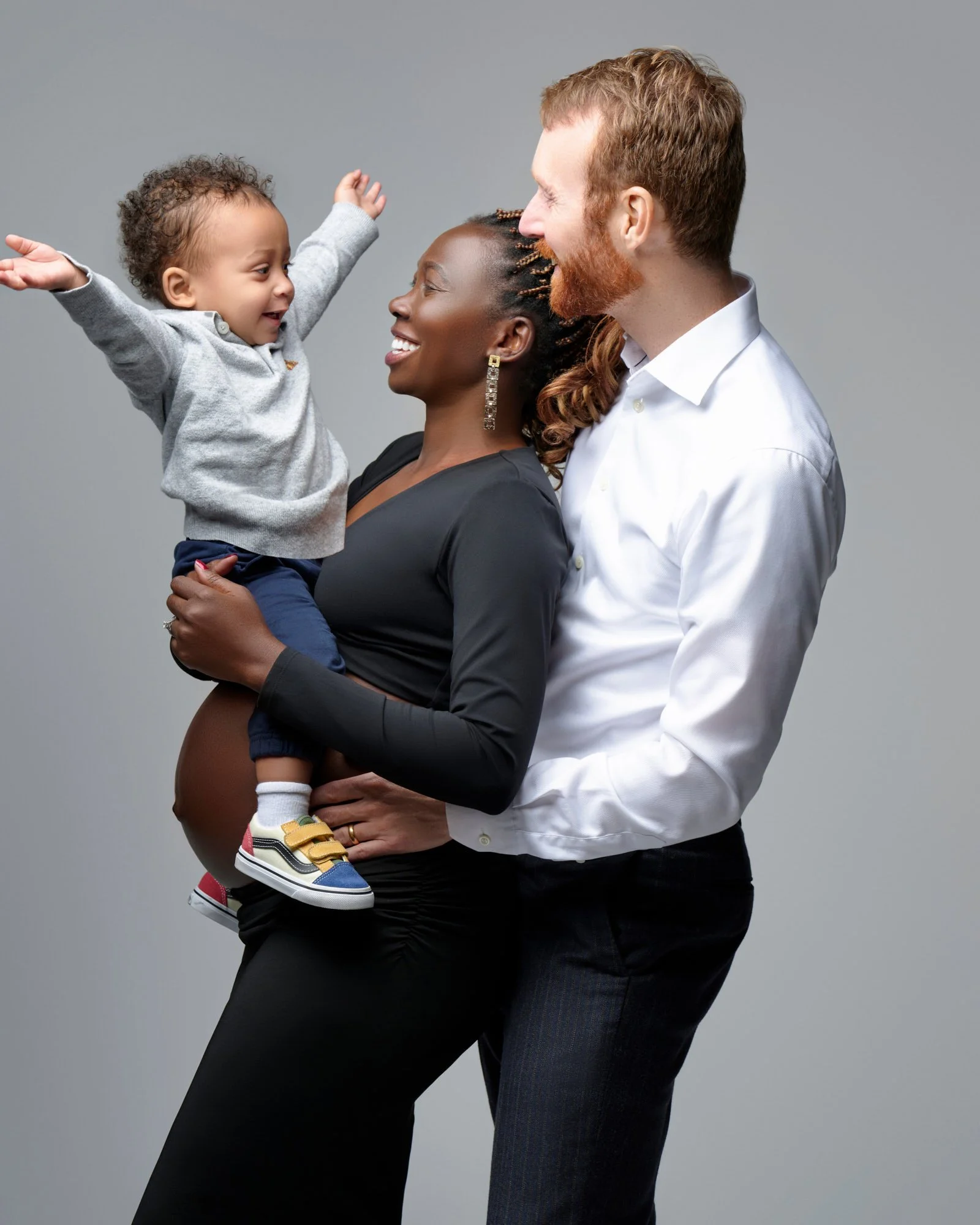 A happy family of three with a pregnant woman holding a child, standing together and smiling at each other against a plain gray backdrop.