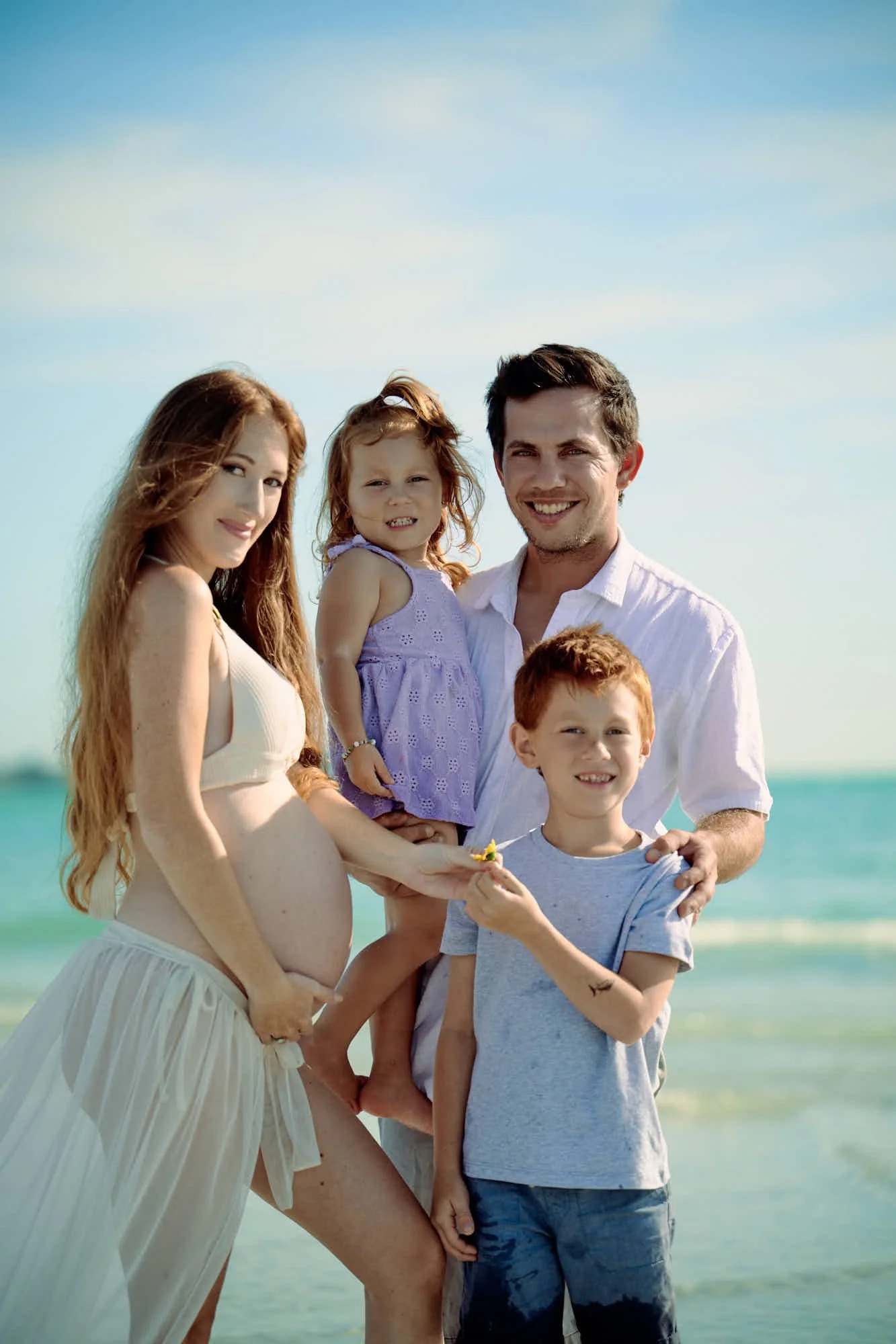 Family of four on a white sand beach, toddler on her hip and turquoise water behind.