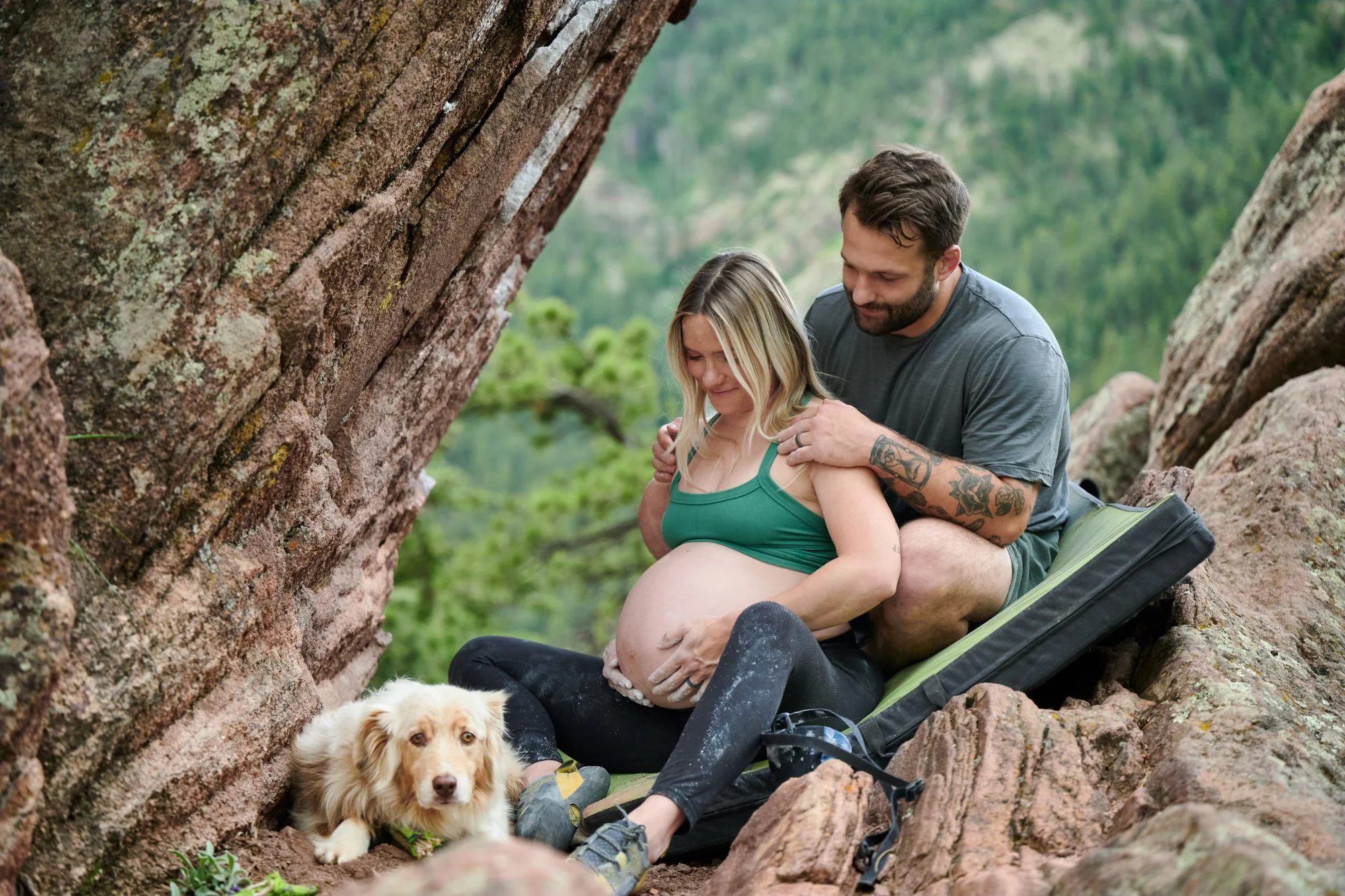 Climbing couple resting on red rock with their dog, his hand on the bump and a crash pad behind.