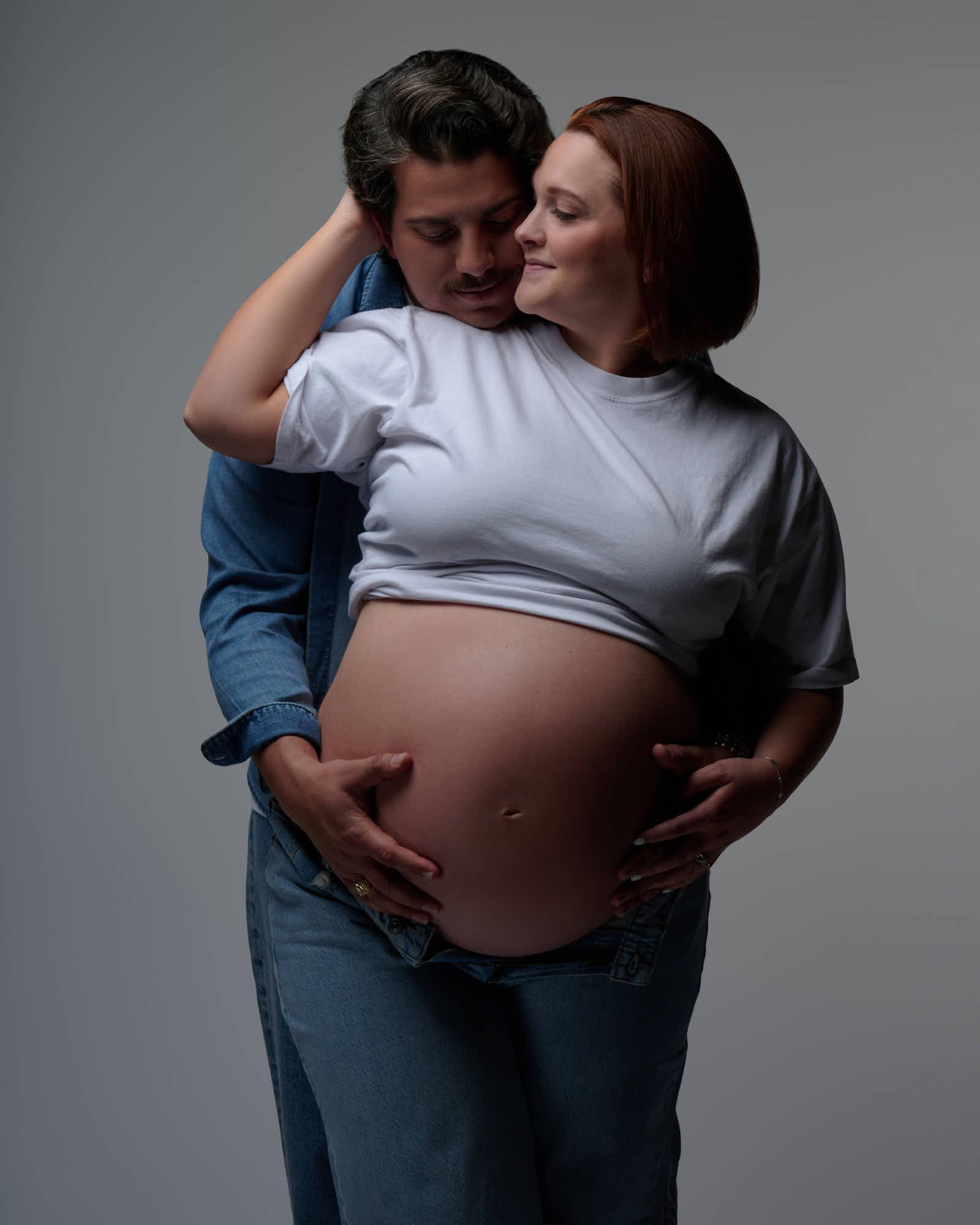 Cheek to cheek in white tees and denim, four hands on the baby bump.