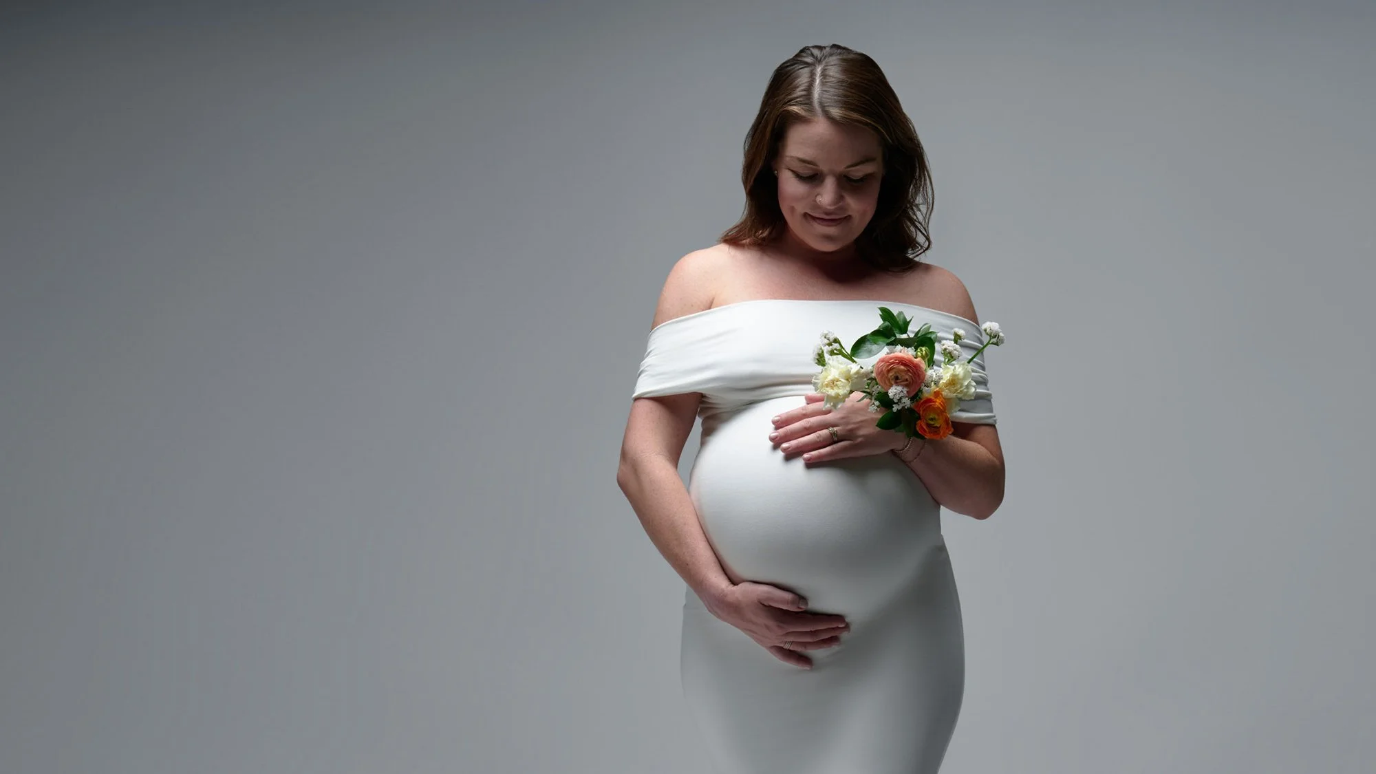 Expecting mother in a white off-shoulder gown holding a bouquet over her bump in the Denver studio.