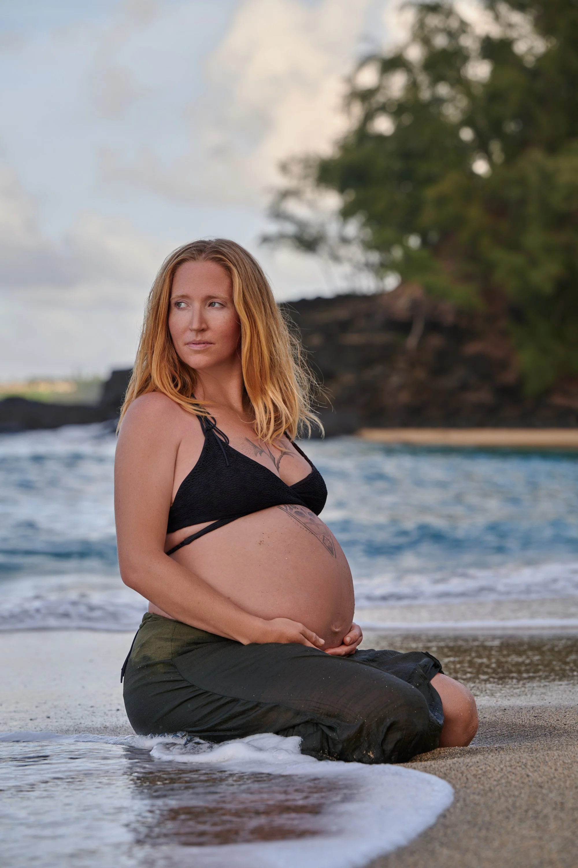Kneeling in the surf in an olive skirt, foam rushing around her on a Kauai beach.