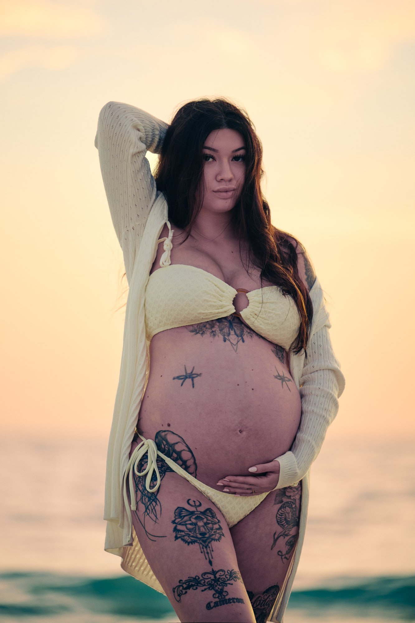 Knit cardigan over a pale yellow two-piece, arm raised, sunset water behind.