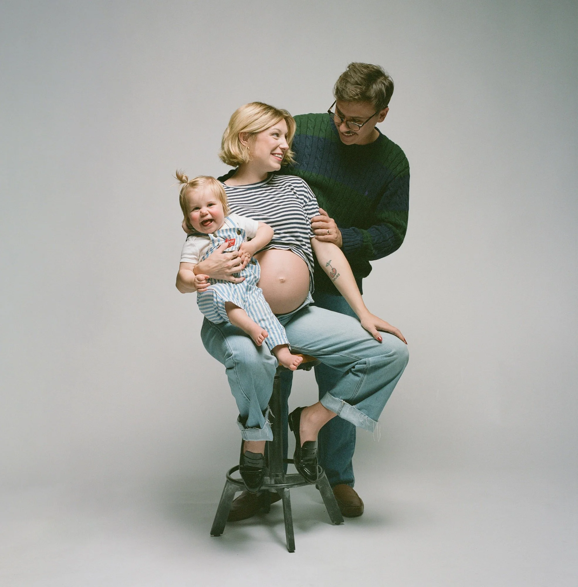 Family of three on a studio stool, toddler on her lap and a bare bump, by Ethan Herrold.