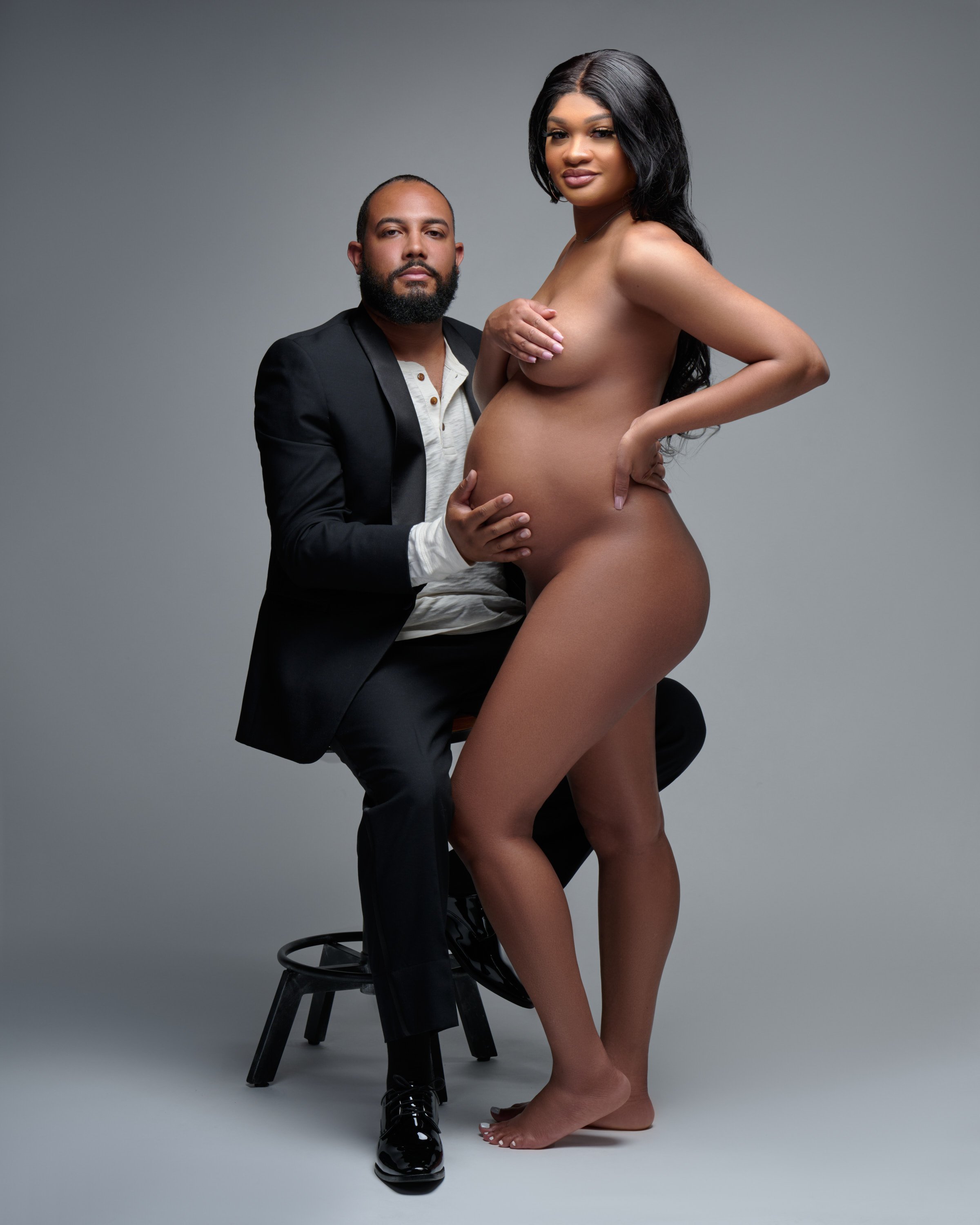 Elegant studio pairing: partner seated in a suit, mother standing with bump.
