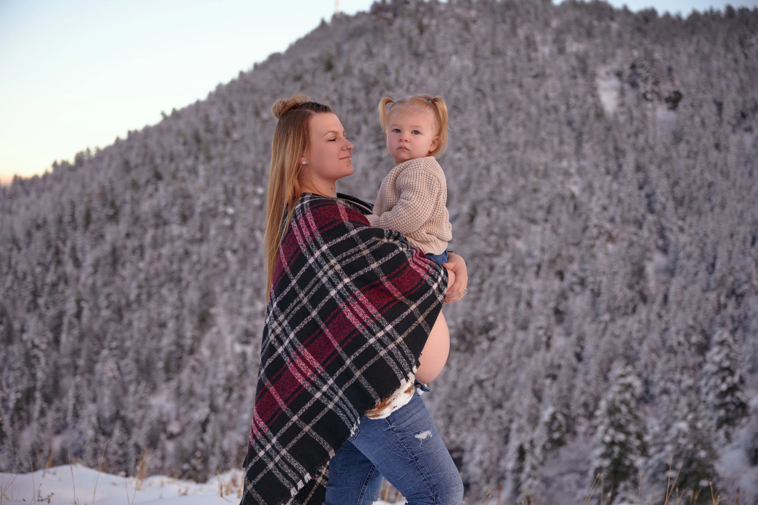 Pregnant woman in a plaid wrap holding her toddler against a snowy mountain backdrop in Evergreen, Colorado