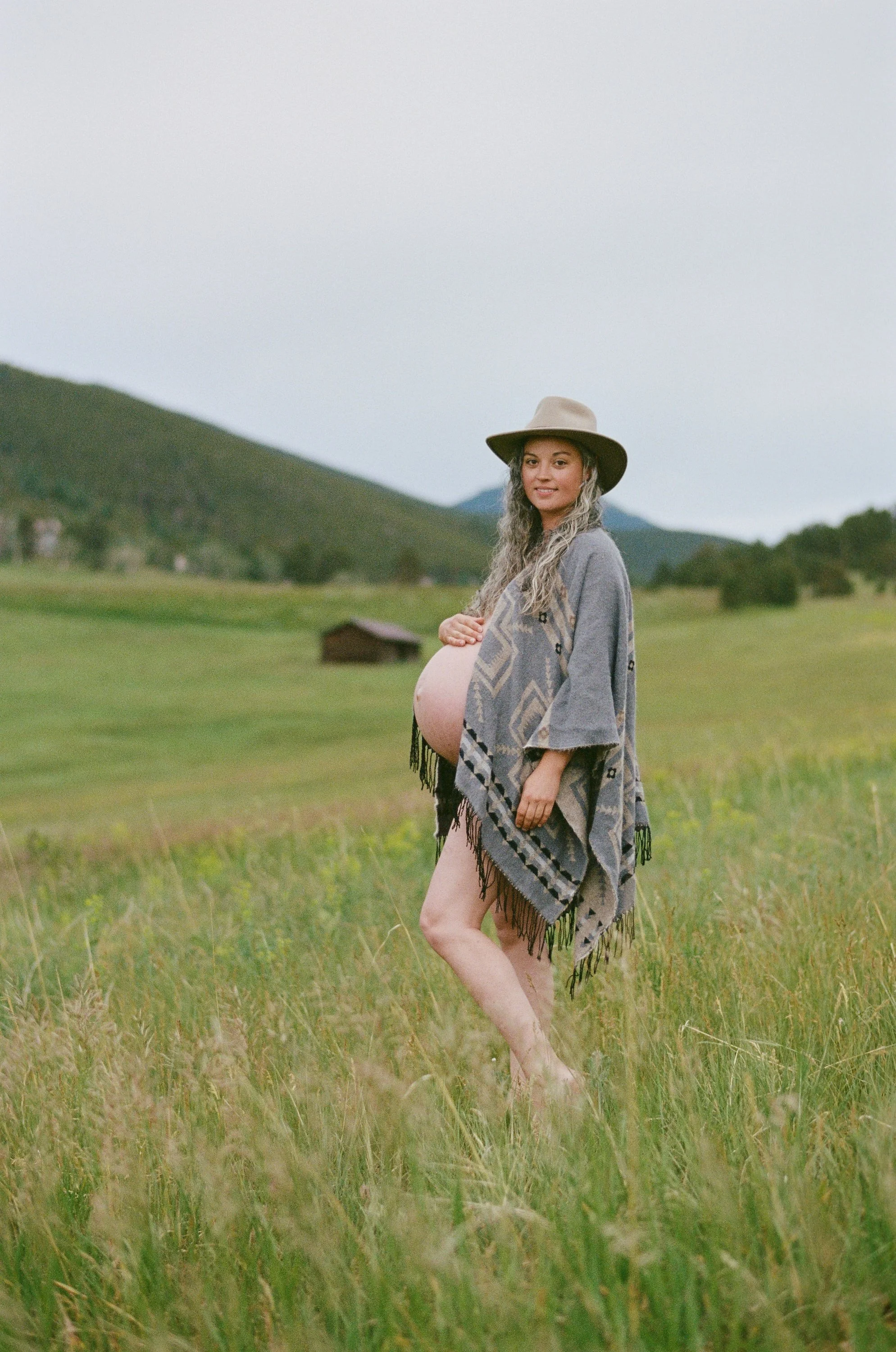 Wide-brim hat and a fringed poncho in tall summer grass, Colorado ranch land rolling out behind.