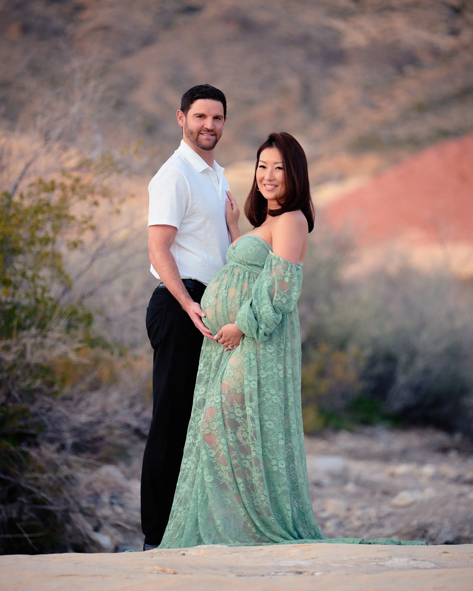 Couple smiling on a sandstone ledge, sage lace gown and desert brush with red cliffs behind.