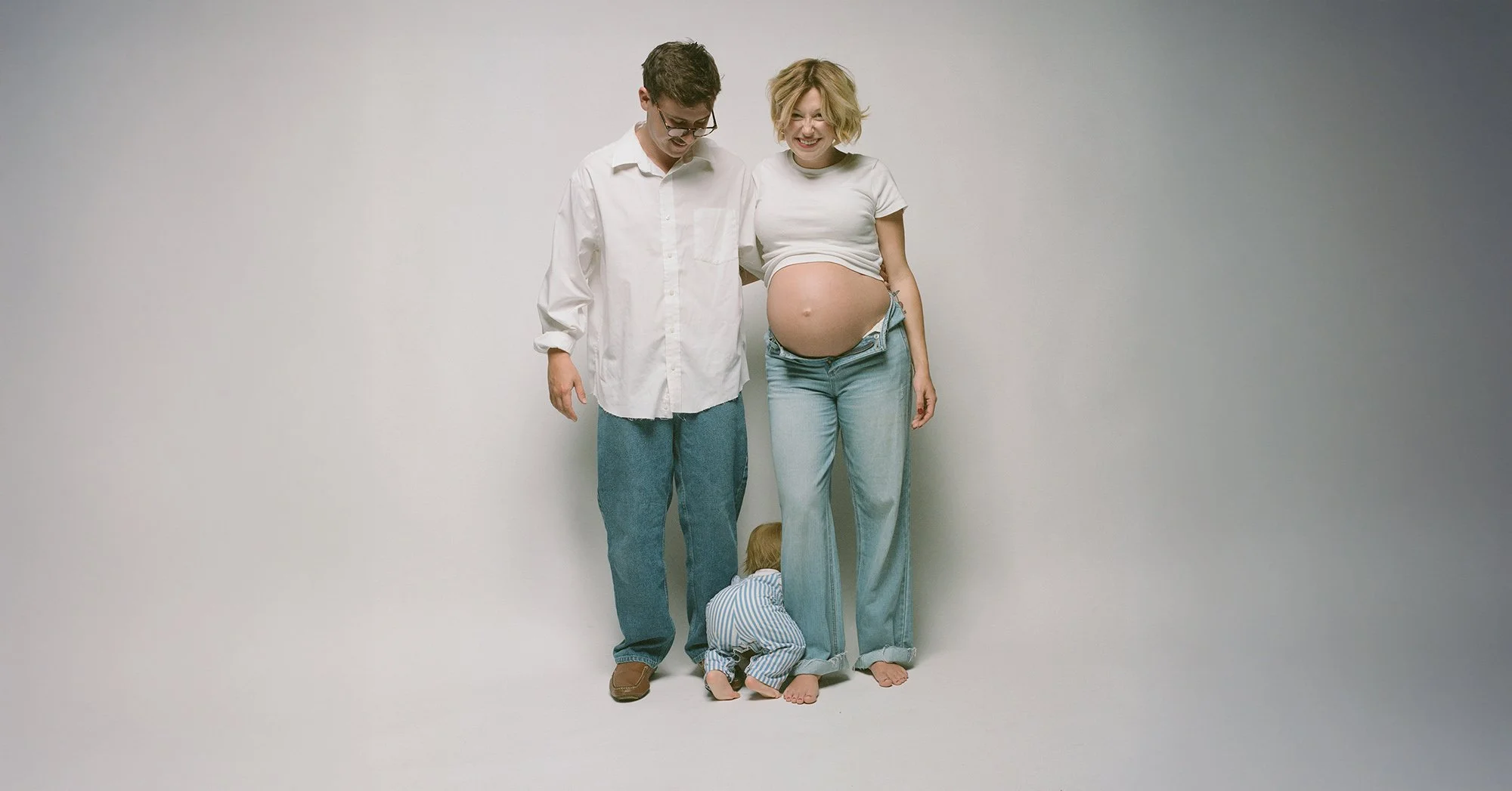 Expecting couple in white tees and jeans laughing in the studio, toddler at their feet.