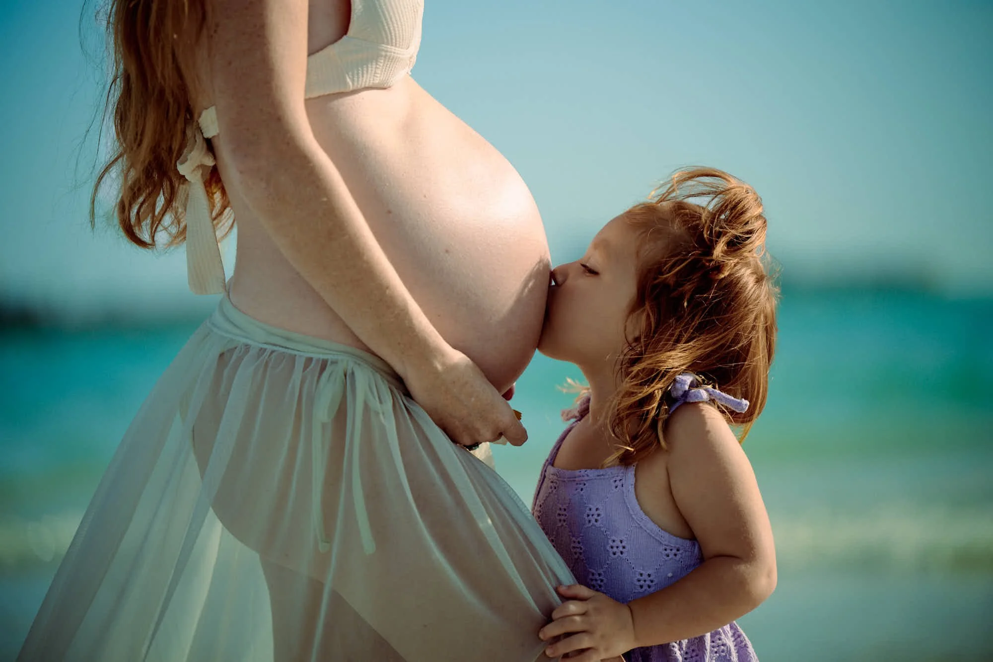 Small daughter kissing the bump on the beach, sheer cover-up lifting in the breeze.