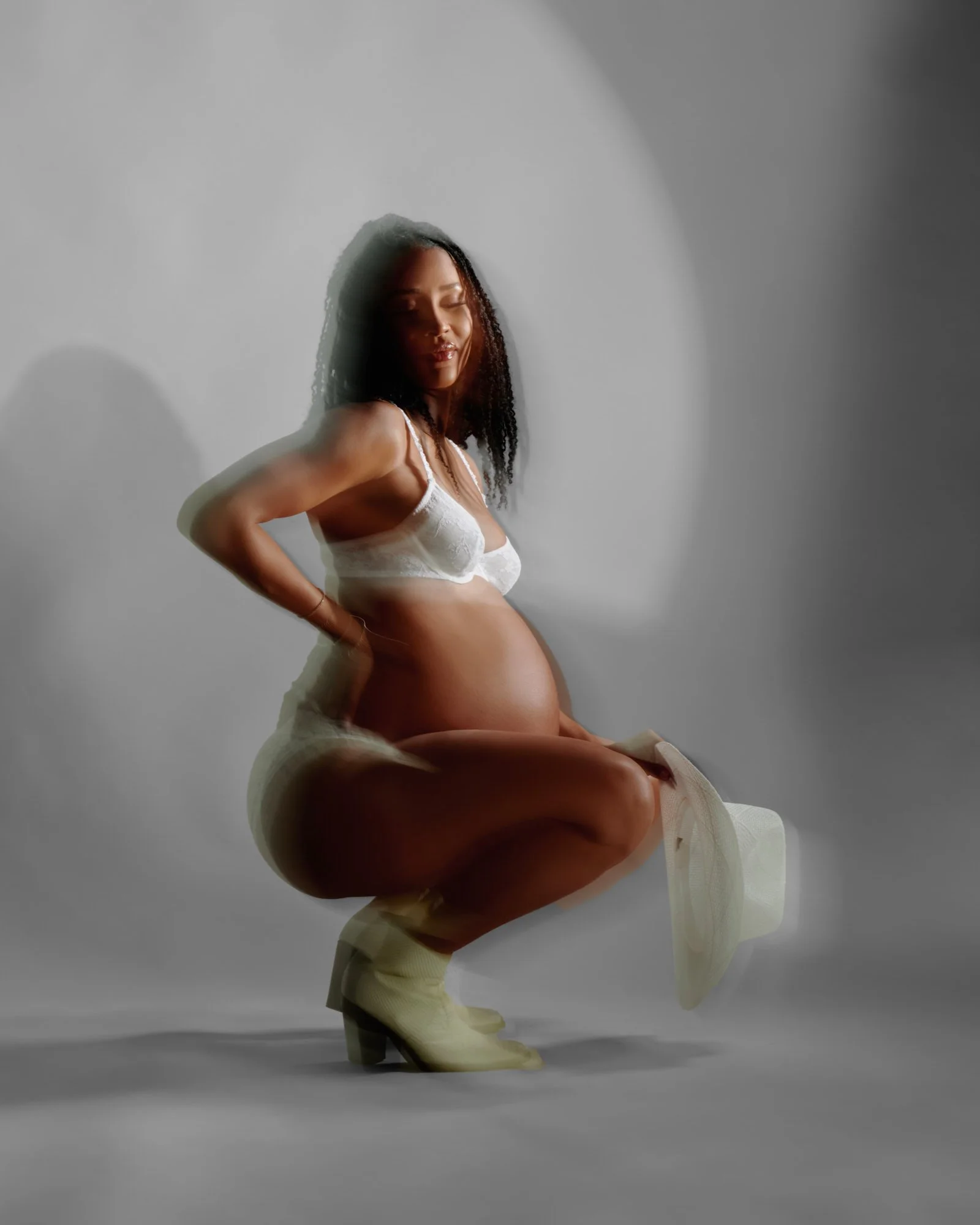 Crouched in white lingerie and cowgirl boots, warm rim light on grey.