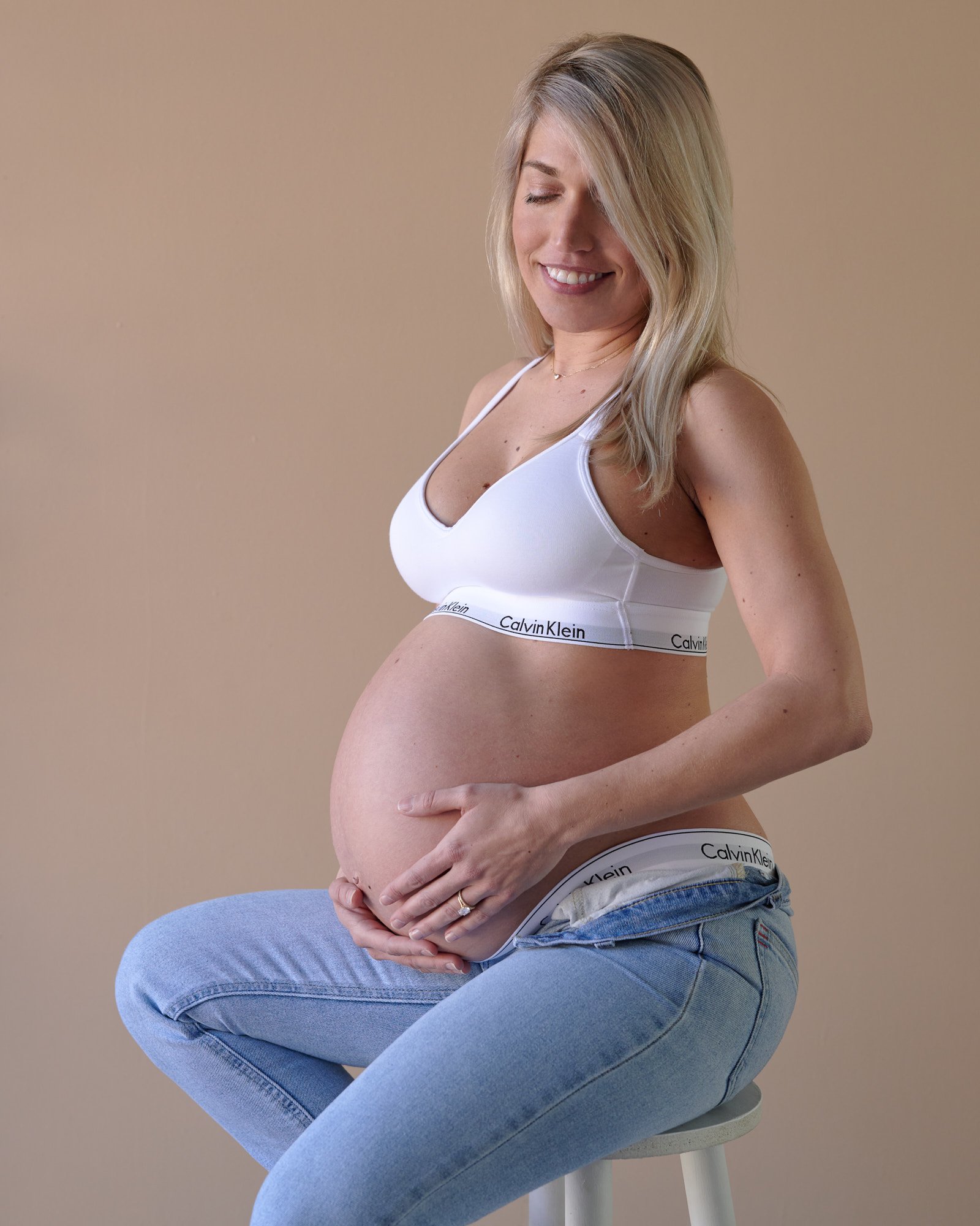 Smiling mother-to-be in a white bralette and unbuttoned jeans seated on a stool, warm tan backdrop.