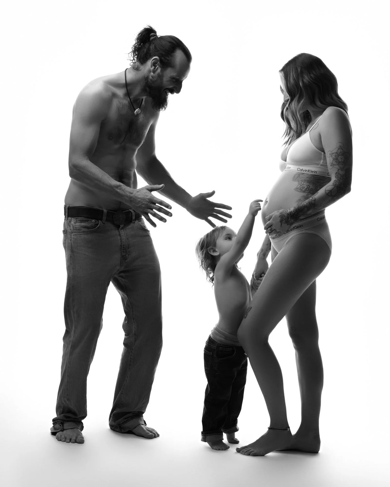 A family portrait of a man, woman, and small child in underwear, smiling and touching the woman's pregnant belly against a white background.
