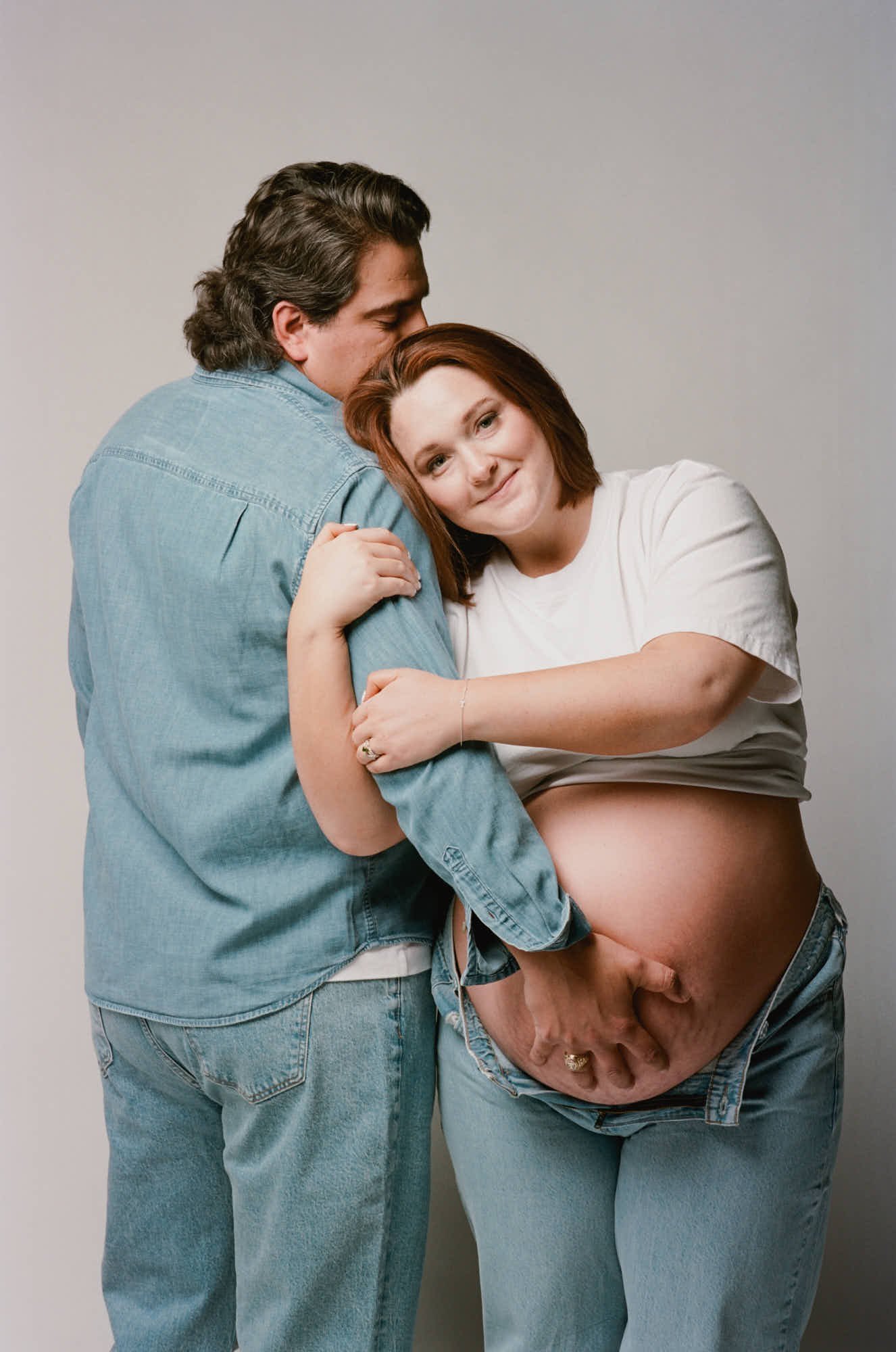 Couple in denim and white cotton, his hands framing her bare bump against a soft studio backdrop.