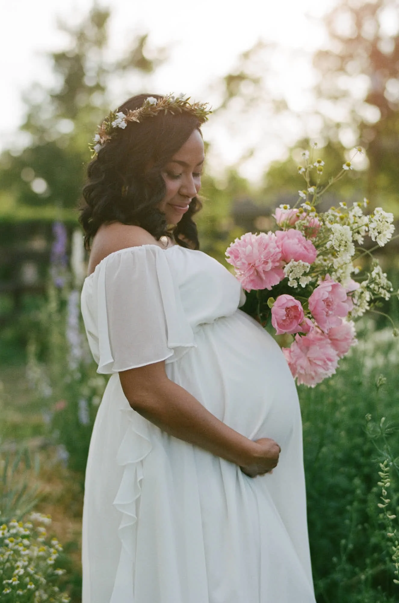 Peonies held low against a white gown, summer green going soft behind her on film.
