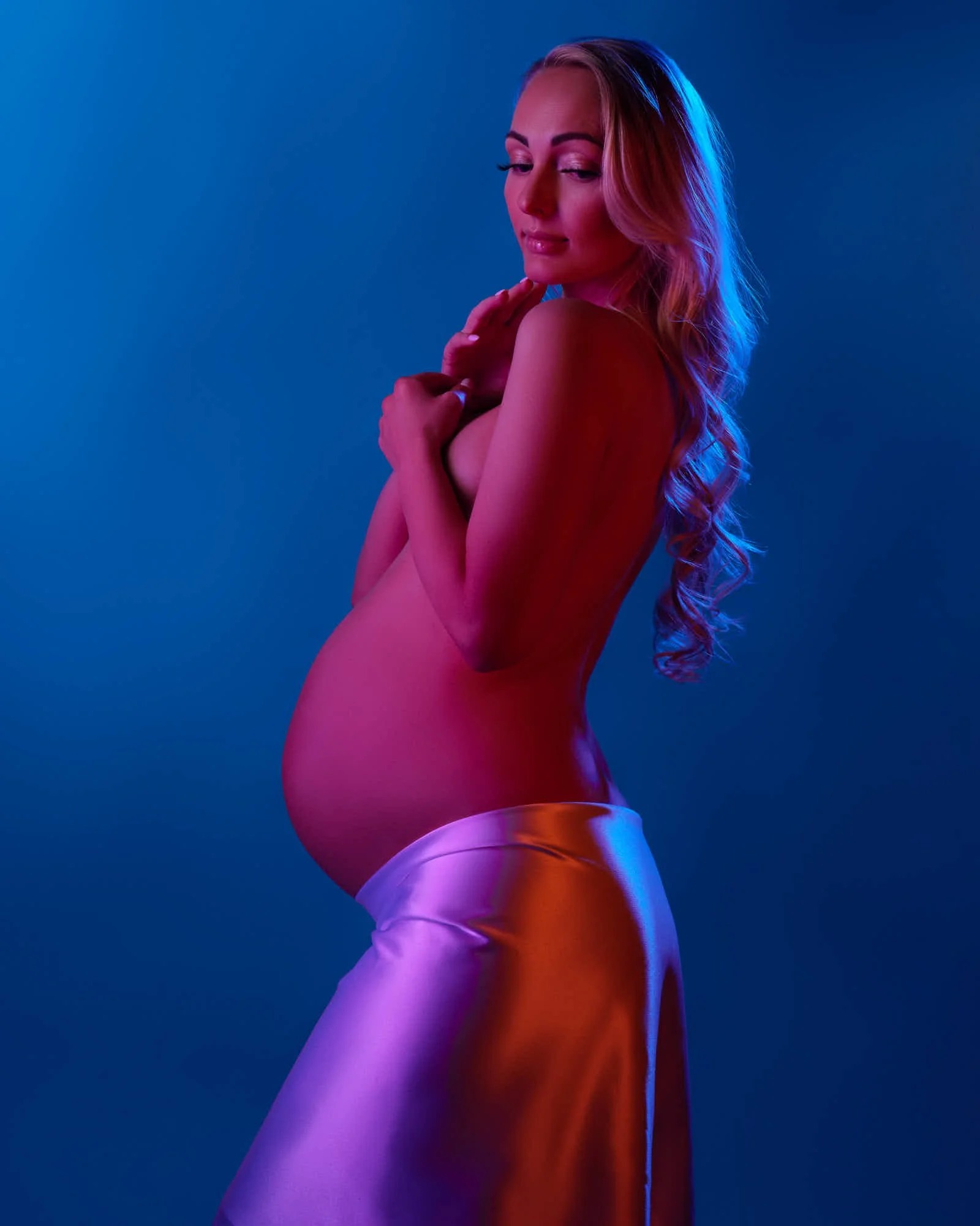 Expecting mother in a lilac satin skirt under blue gel light with a warm rim, chin to shoulder.