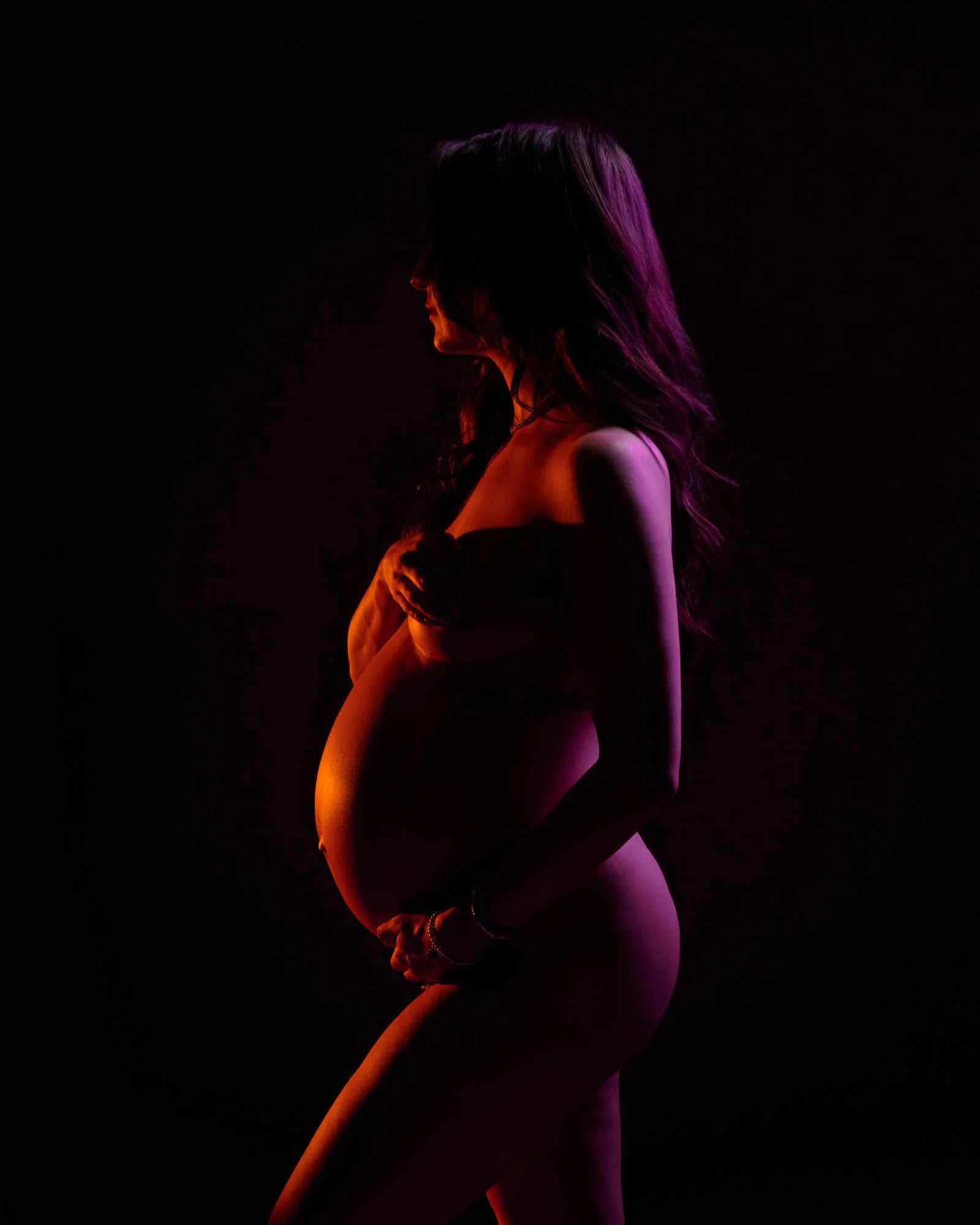 Pregnant woman in profile under orange and pink gel lighting, cradling her bump against a black backdrop.