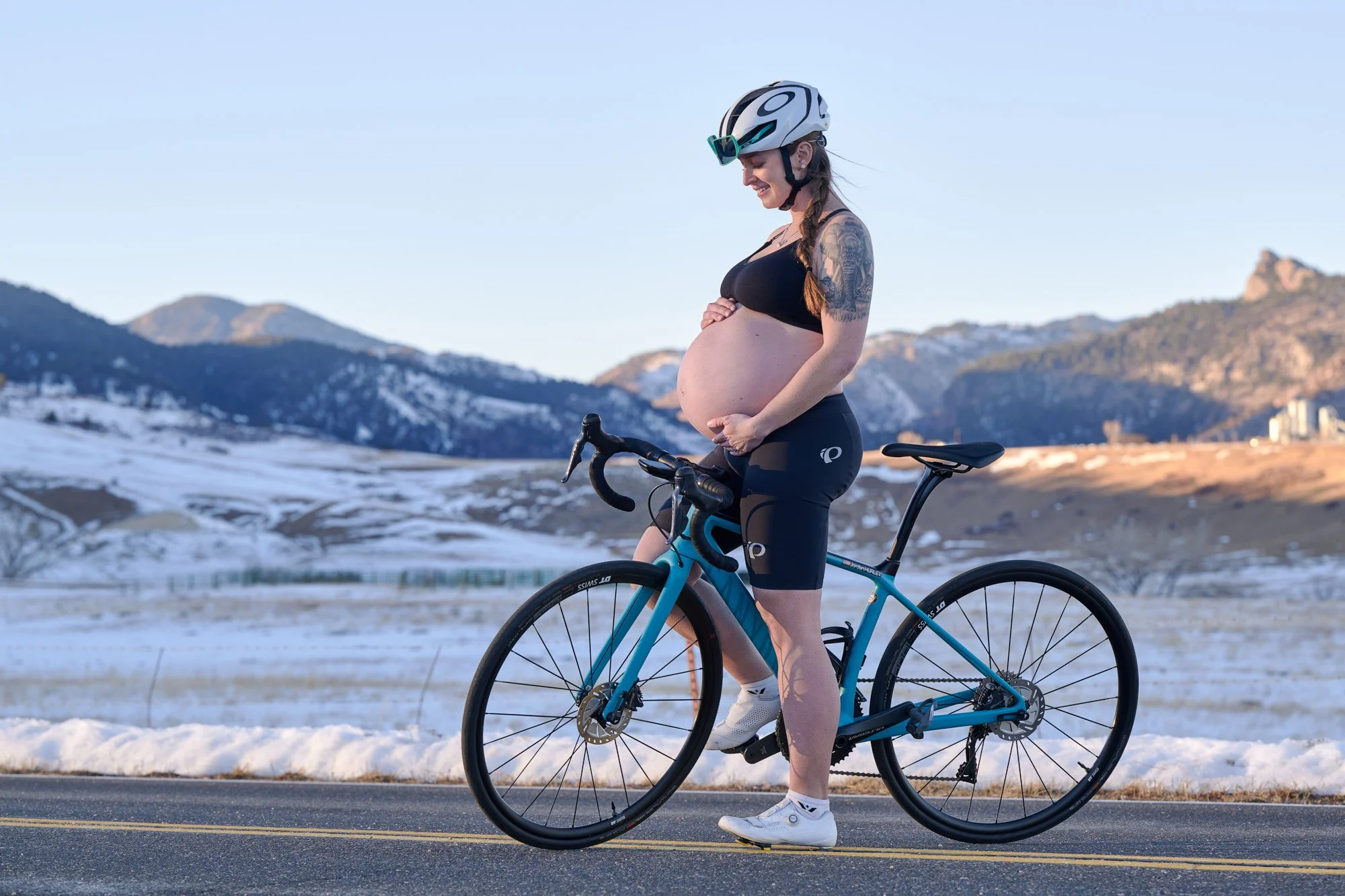 Beside the road bike with snowy peaks behind, both hands cradling the bump.