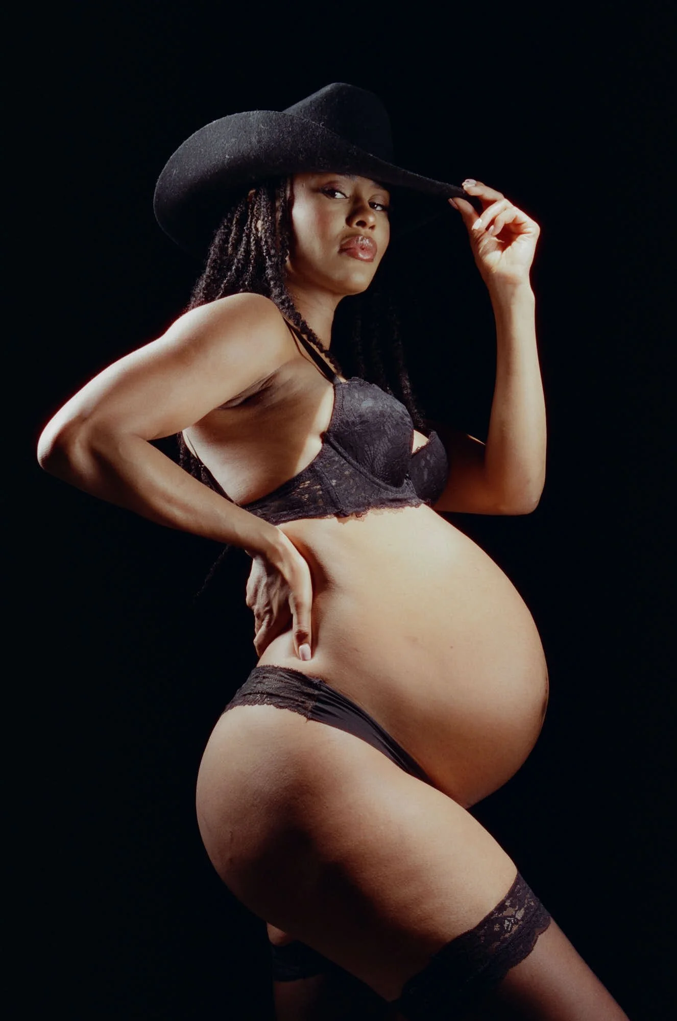 Black hat brim in hand, lace and stockings against a dark wall, one hip pushed forward.