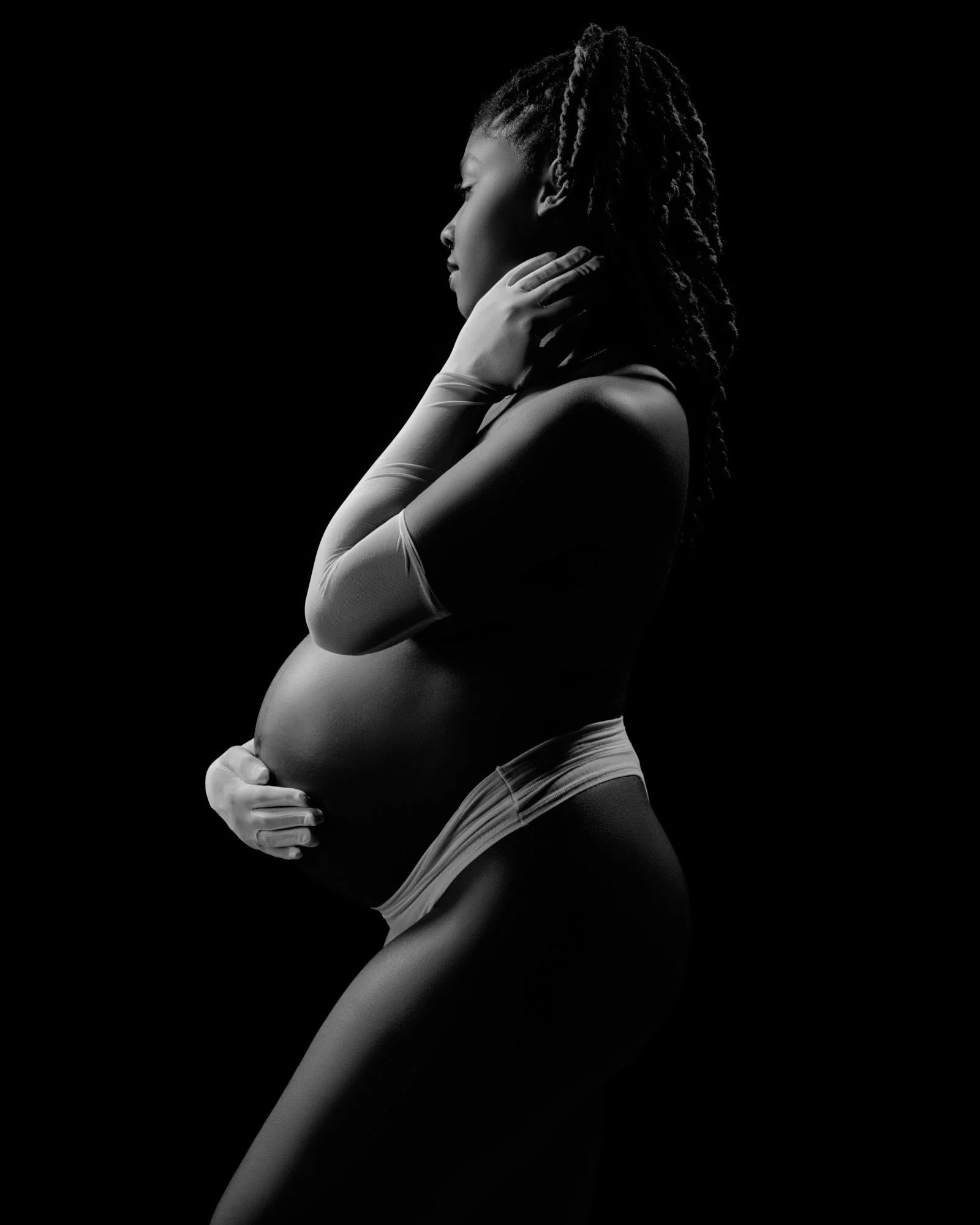 Low-key side light and locs, arms folded in a quiet powerful pose.