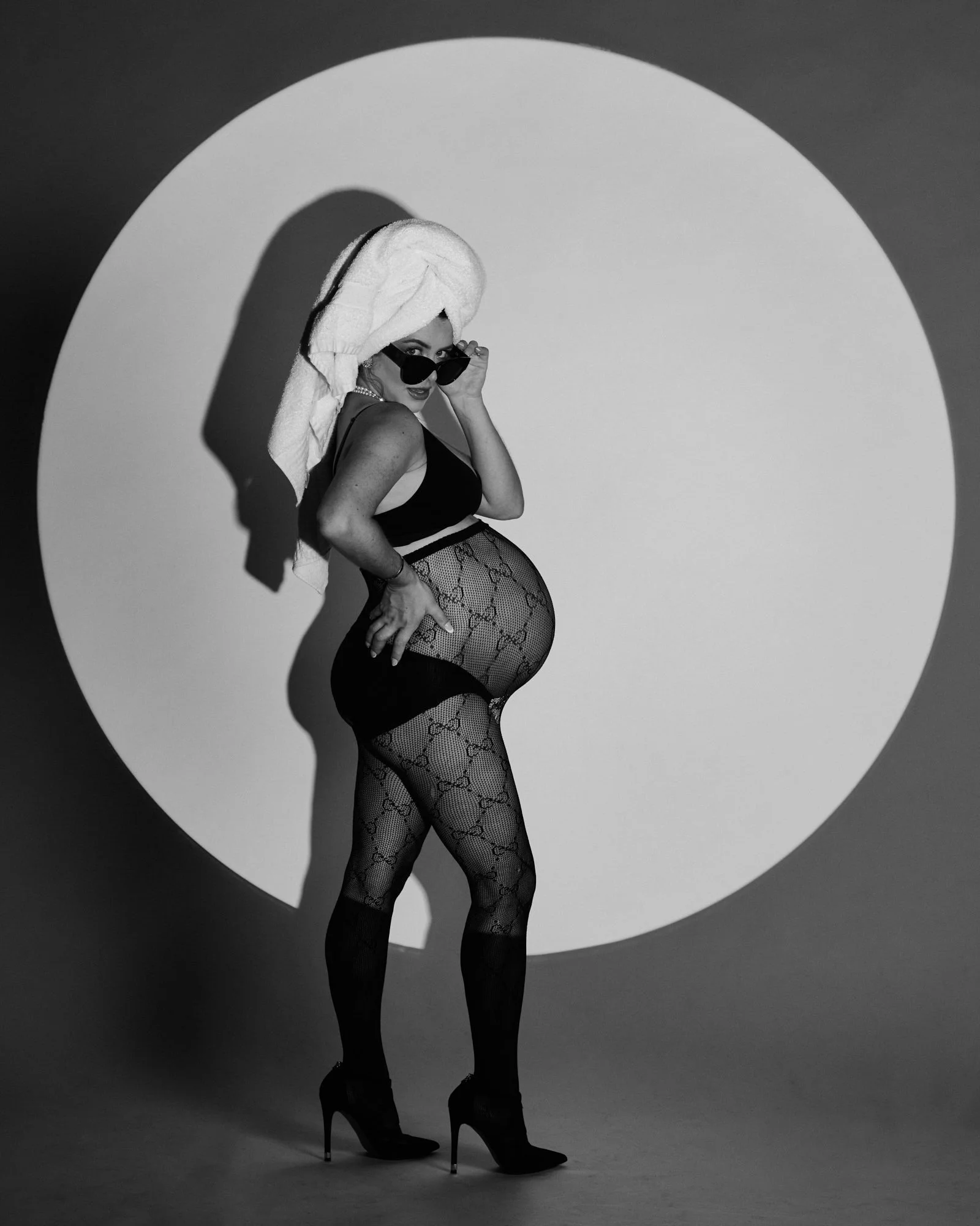 Black-and-white photo of a woman in lingerie and fishnet stockings, with a towel on her head, posing with sunglasses, standing in a circle of light on the wall.