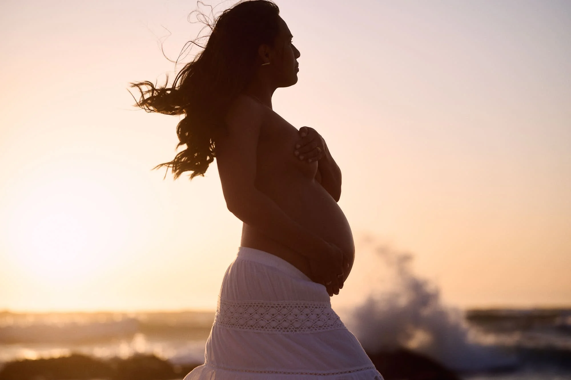 Backlit at sunset with hands on her bump, a wave breaking white against the rocks behind.